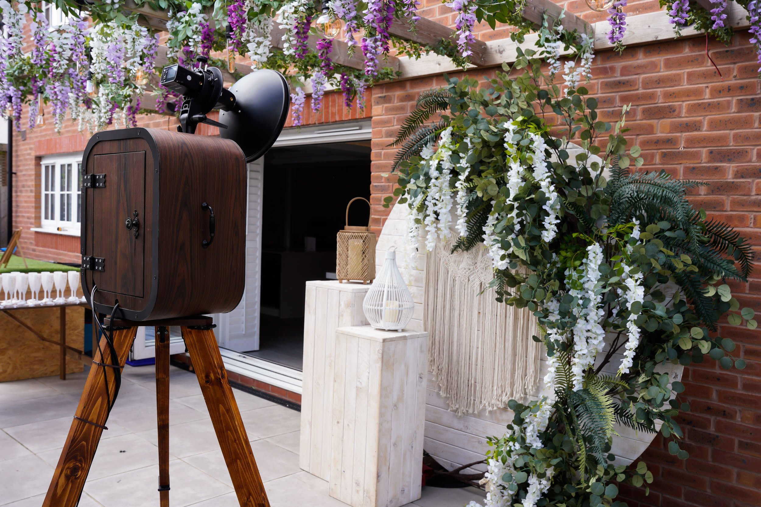 Decorated outdoor space with a cascading floral arrangement, a wooden tripod-mounted camera, and various decorative items on white stands, set against a brick wall.