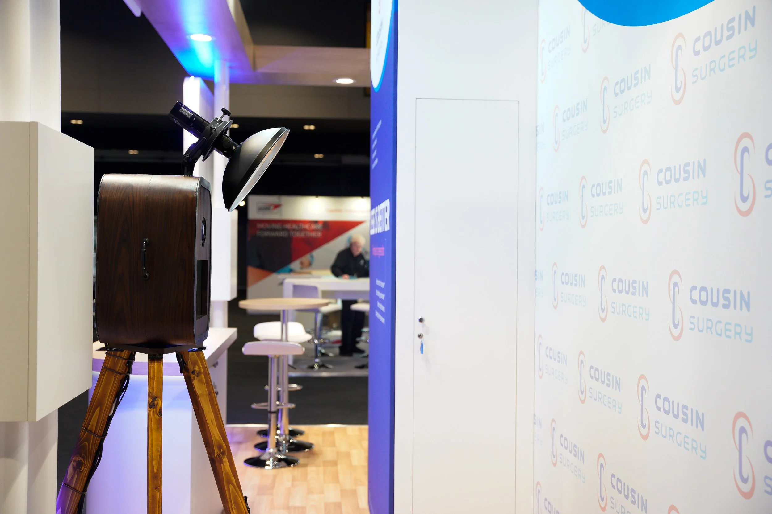 Photo of a camera on a tripod in front of a blue and white backdrop with the text "Cousin Surgery." A person is sitting at a table in the background.