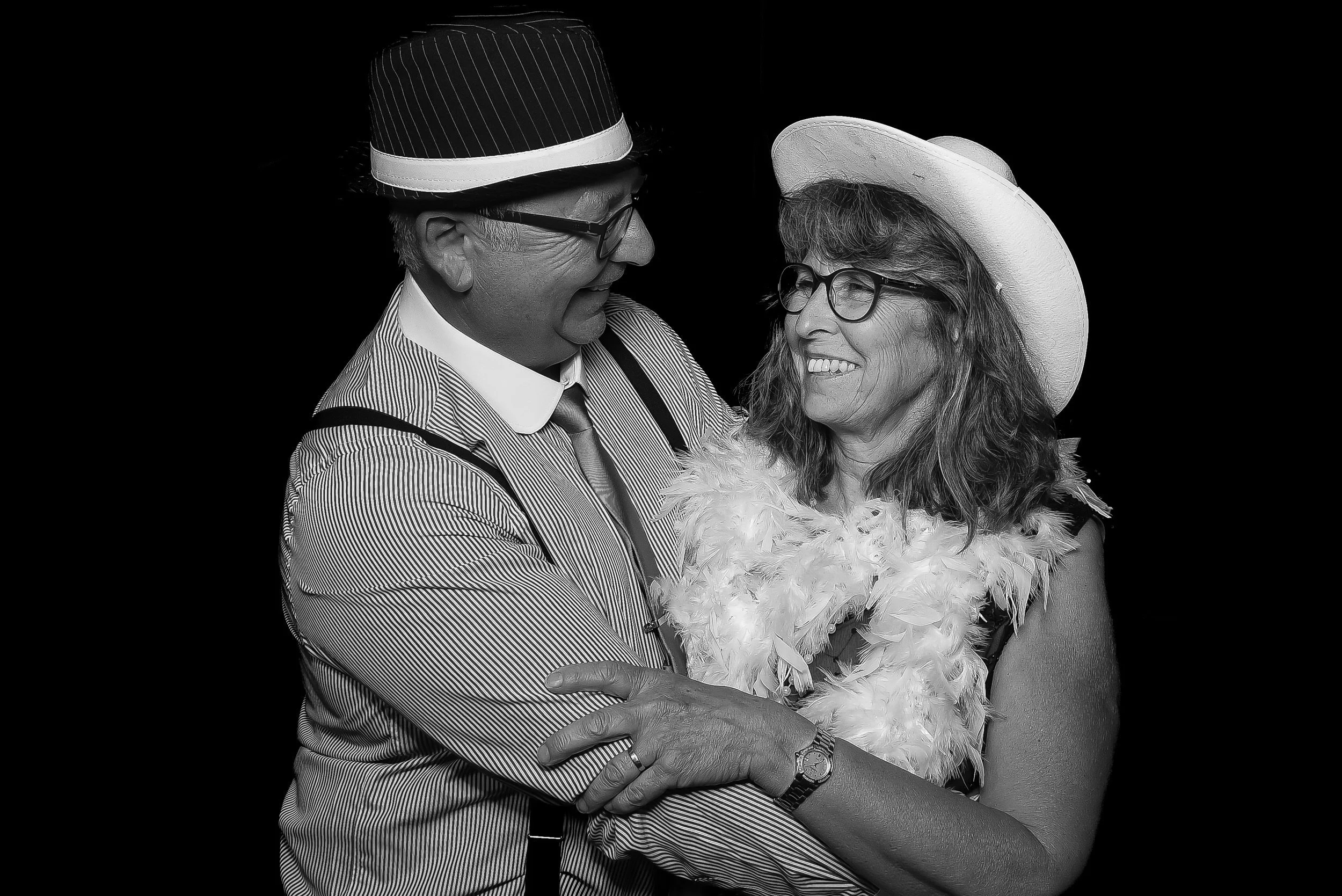 A happy man and woman smiling and looking at each other, embracing. The man is wearing a pinstripe shirt, tie, suspenders, glasses, and a black hat with a white band. The woman is wearing glasses, a large brimmed hat, and a feathered boa.