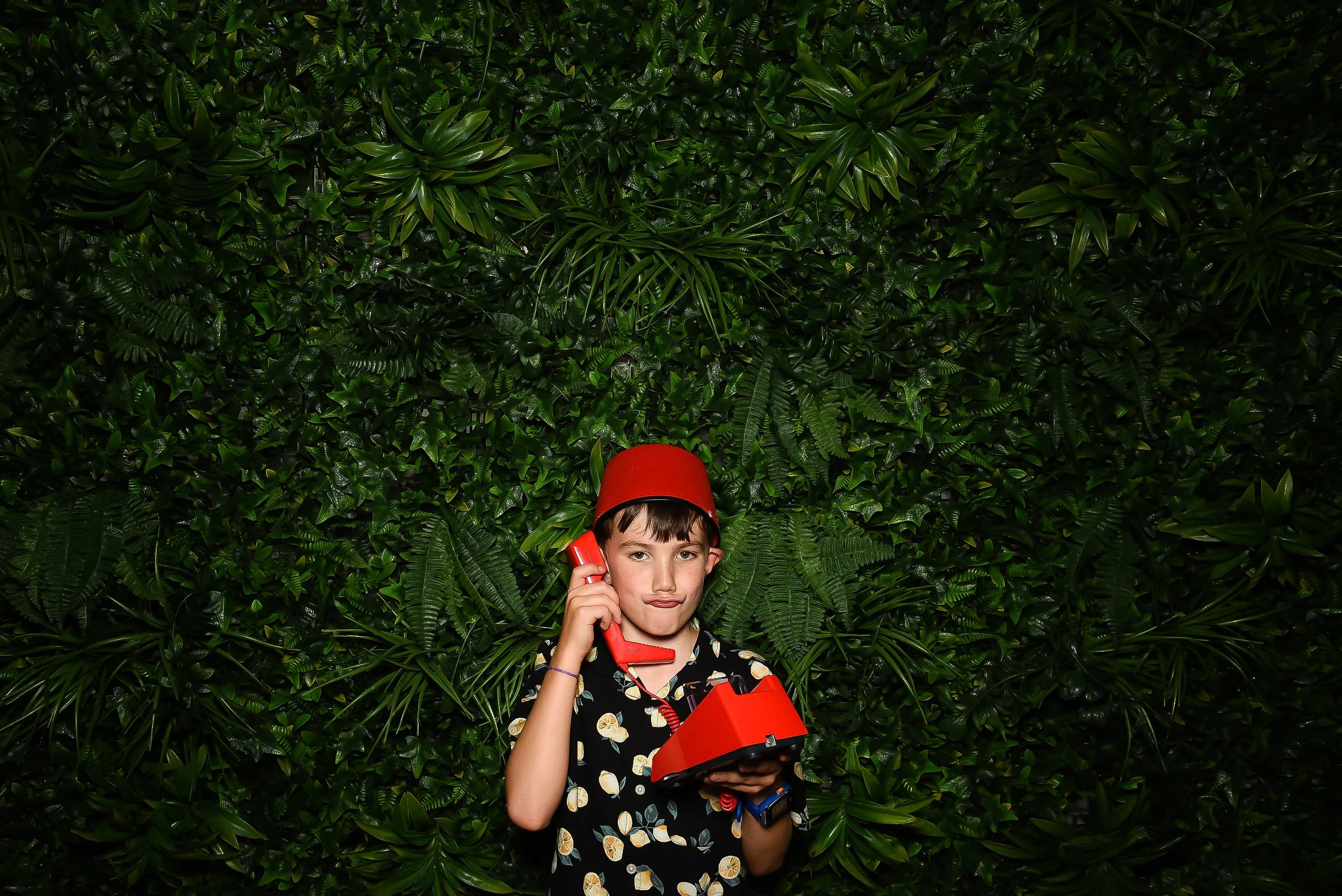 A boy wearing a black shirt with a lemon print, holding a red vintage rotary phone to his ear, and a red bucket hat, standing in front of a lush green leafy wall.