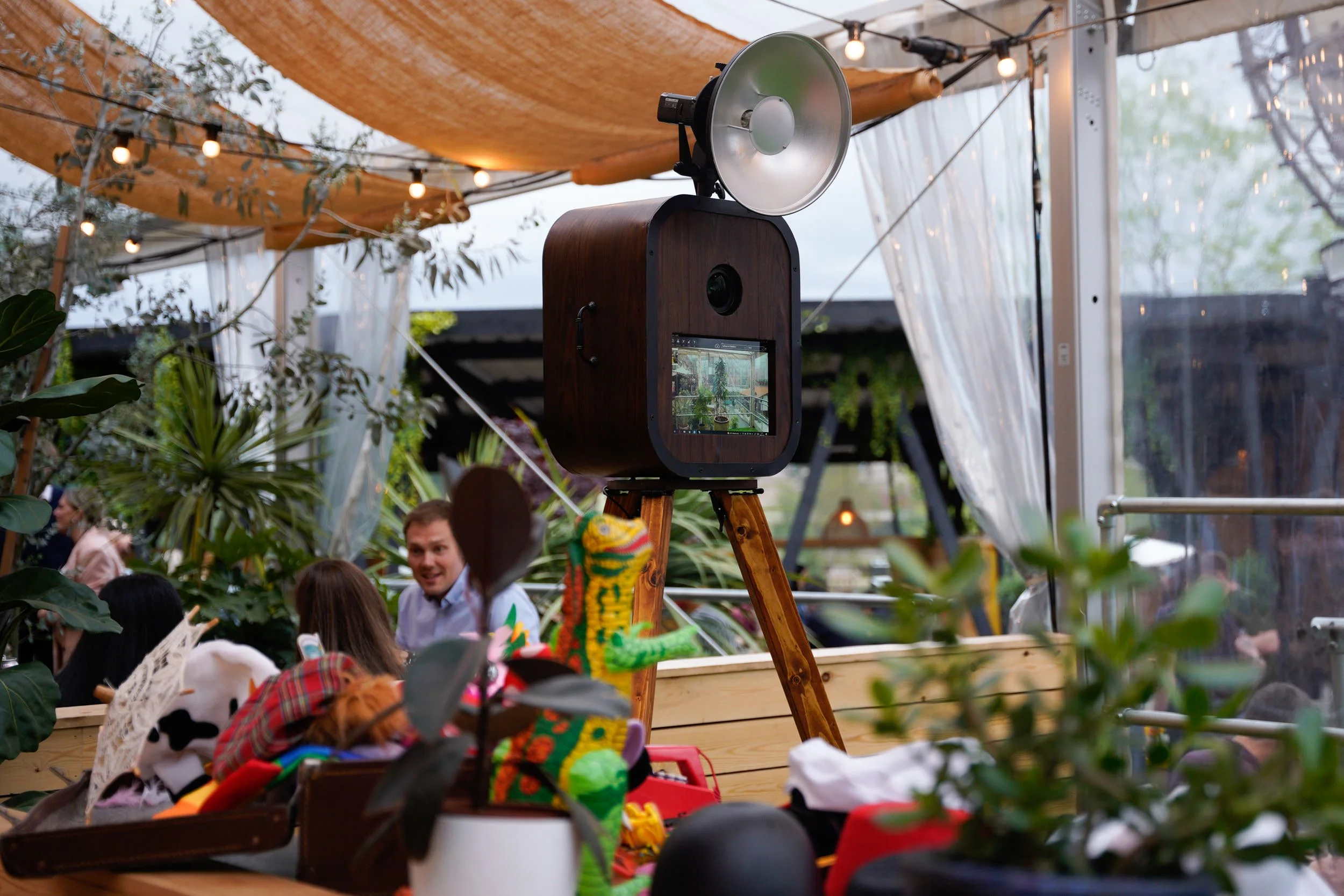 A photo of an outdoor or semi-outdoor space with a large vintage style camera on a tripod, surrounded by people, plants, and string lights.