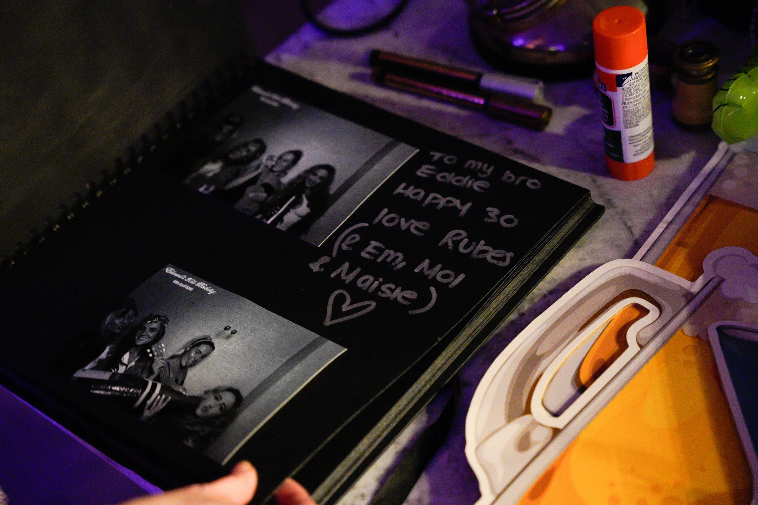 A scrapbook or photo album on a table with black and white photographs of young women, a message written on a black page reading 'To my bro Eddie, happy 30 love Rubes (E Emmanuel) & Naise', a heart symbol, and various craft supplies like glue stick, 