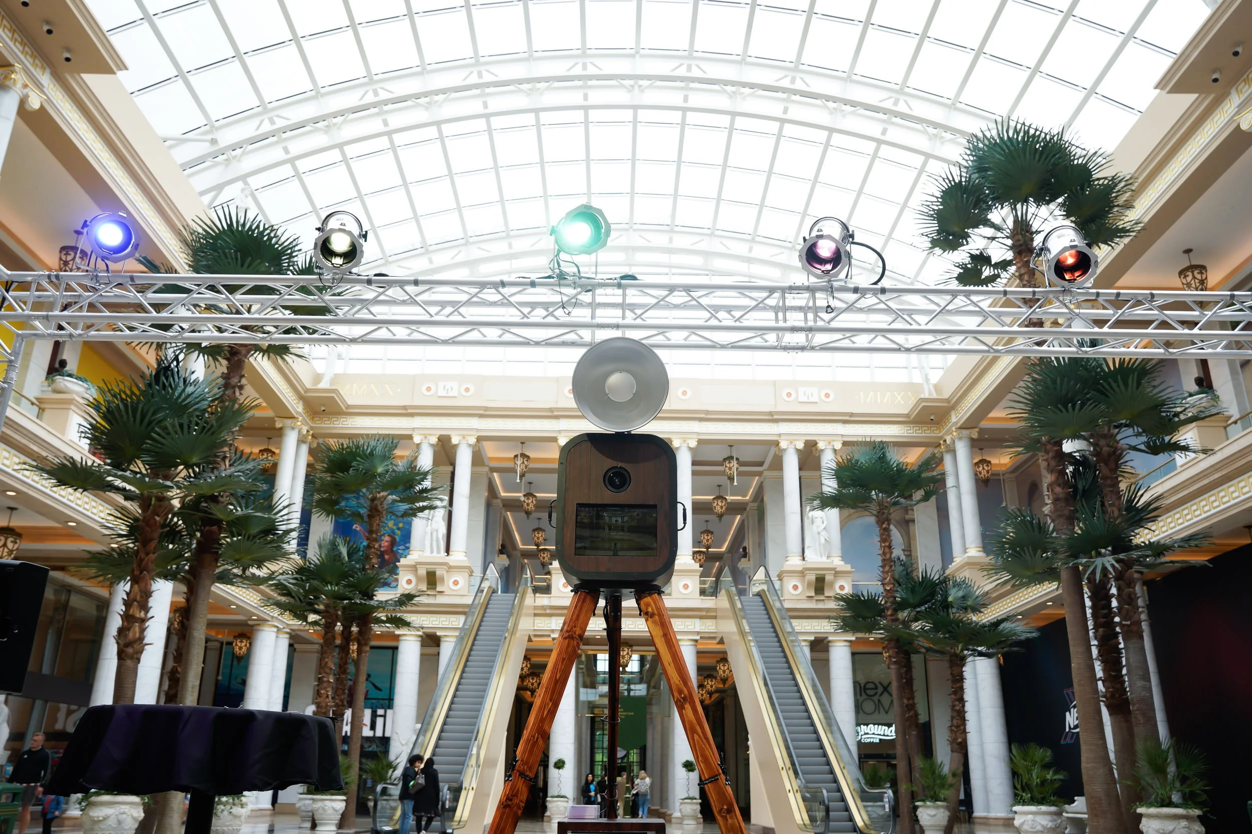 A large camera with a speaker on top, mounted on a tripod, in a shopping mall with palm trees, escalators, and a glass ceiling.