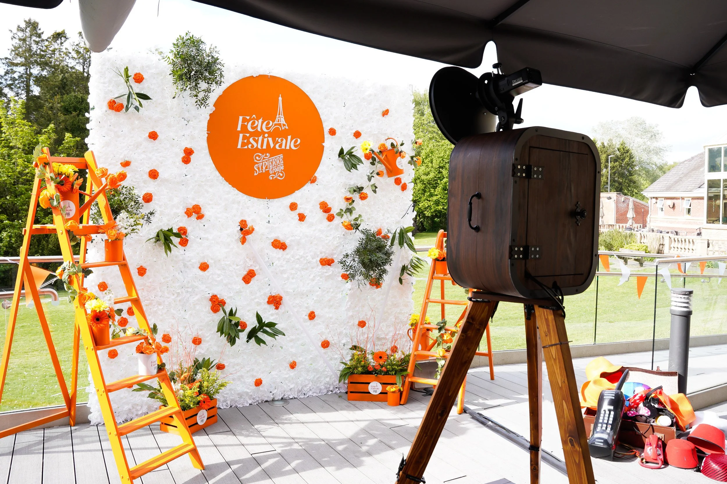 Photo of an outdoor event setup with a white floral backdrop decorated with orange flowers, green foliage, and a large orange circular sign that reads "Fête Estivale" with a graphic of the Eiffel Tower. There are yellow and orange ladders with floral