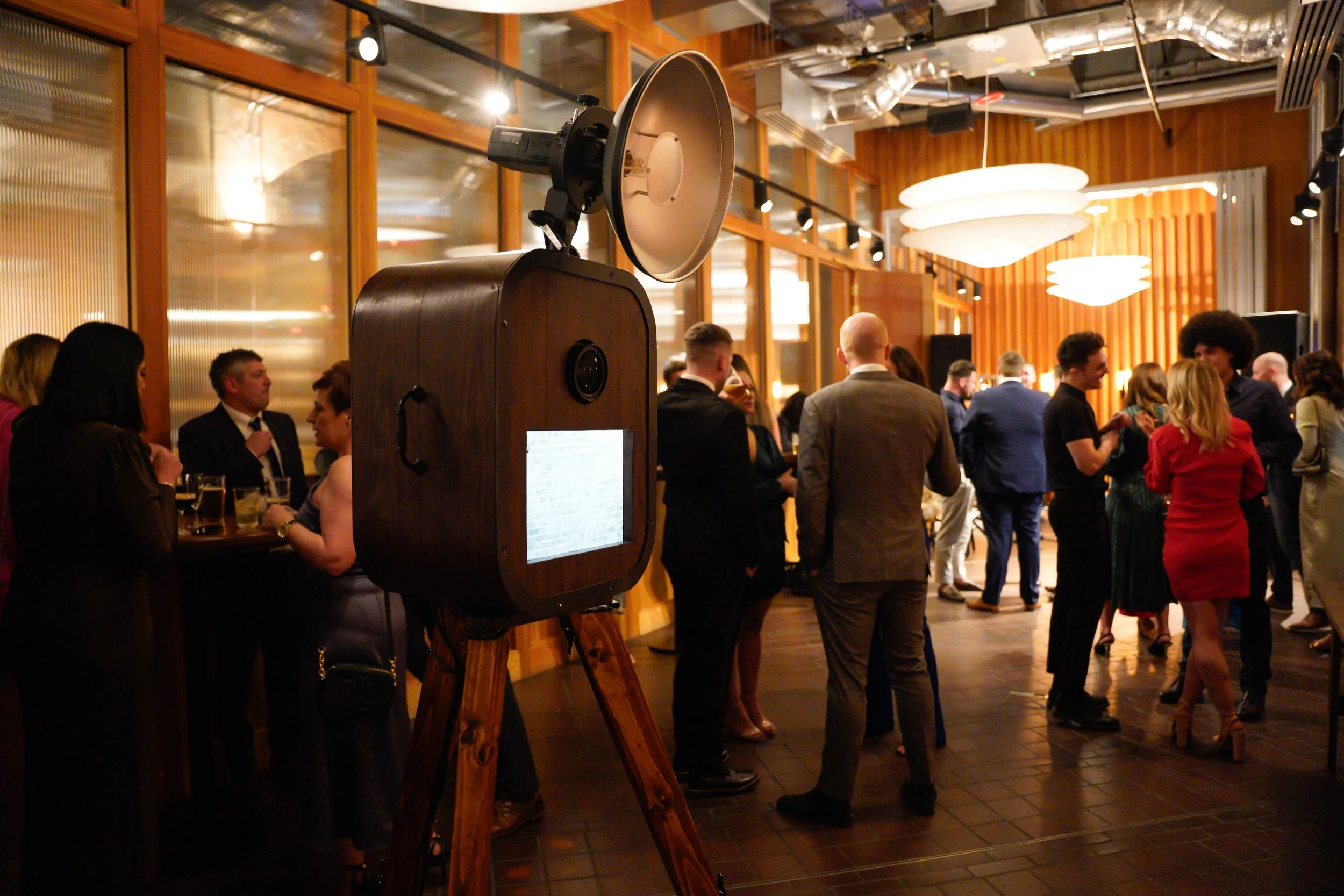 People socializing at a party or reception in a modern, warmly lit venue with large glass windows, wooden walls, and decorative lighting. A vintage camera on a tripod is in the foreground.