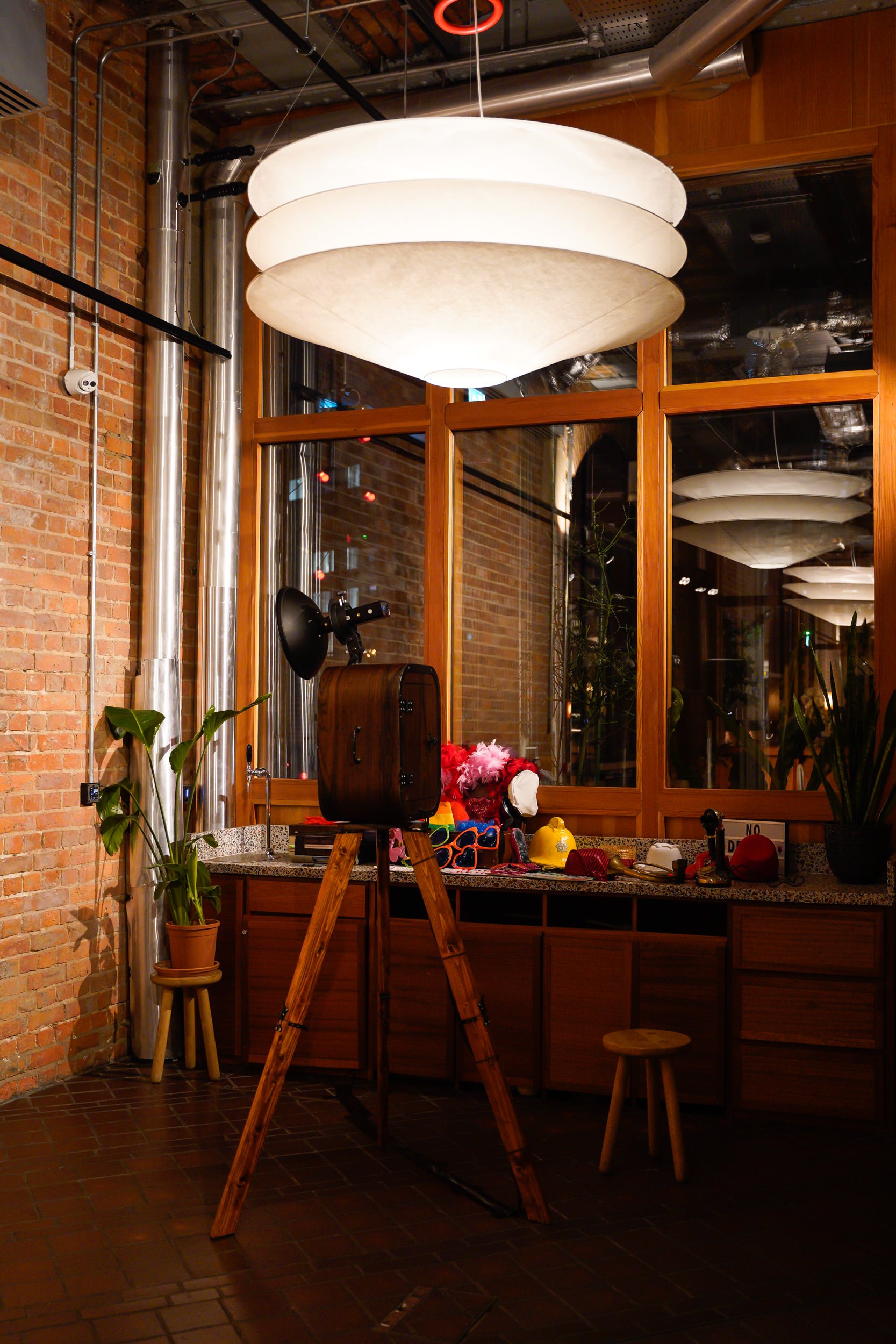 Interior of a room with exposed brick walls, large hanging white lights, a vintage camera on a wooden tripod, and various props and decorations on a counter near a large glass window.