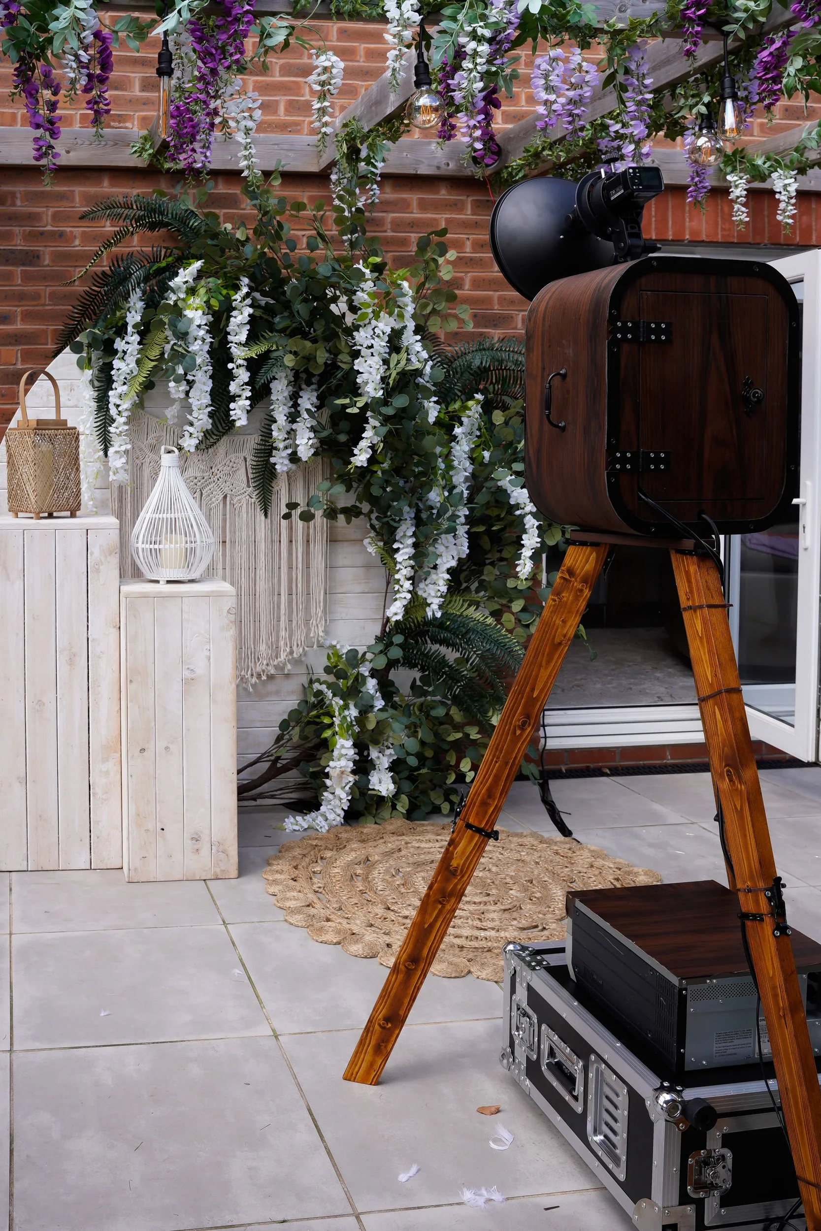 An outdoor photography setup featuring a vintage camera on a tripod, with decorative white, purple, and green floral arrangements, hanging lights, and a brick wall in the background.