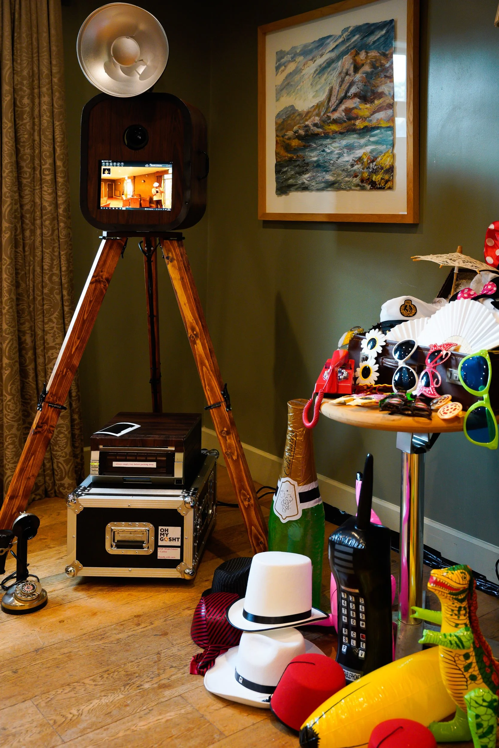 A room corner with a vintage photo booth, a framed landscape painting, and a table with colorful accessories, hats, sunglasses, and inflatable toys. The photo booth has a monitor displaying the interior, and a large studio light is positioned above i