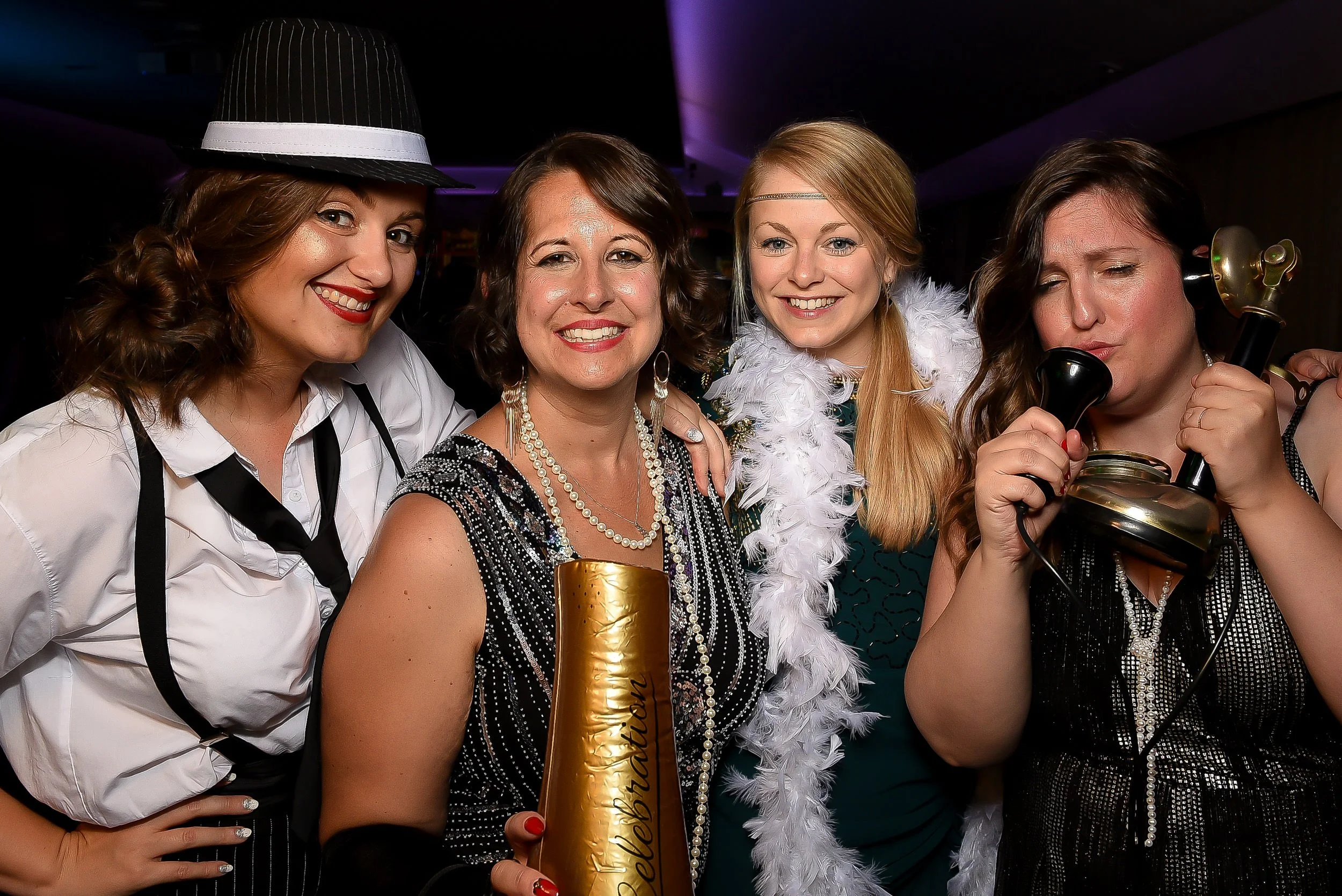 Group of four women at a celebration, dressed in festive clothing, with accessories like pearls and a feather boa, one woman holding a gold party horn, another woman speaking into a vintage telephone, all smiling and enjoying a party environment.