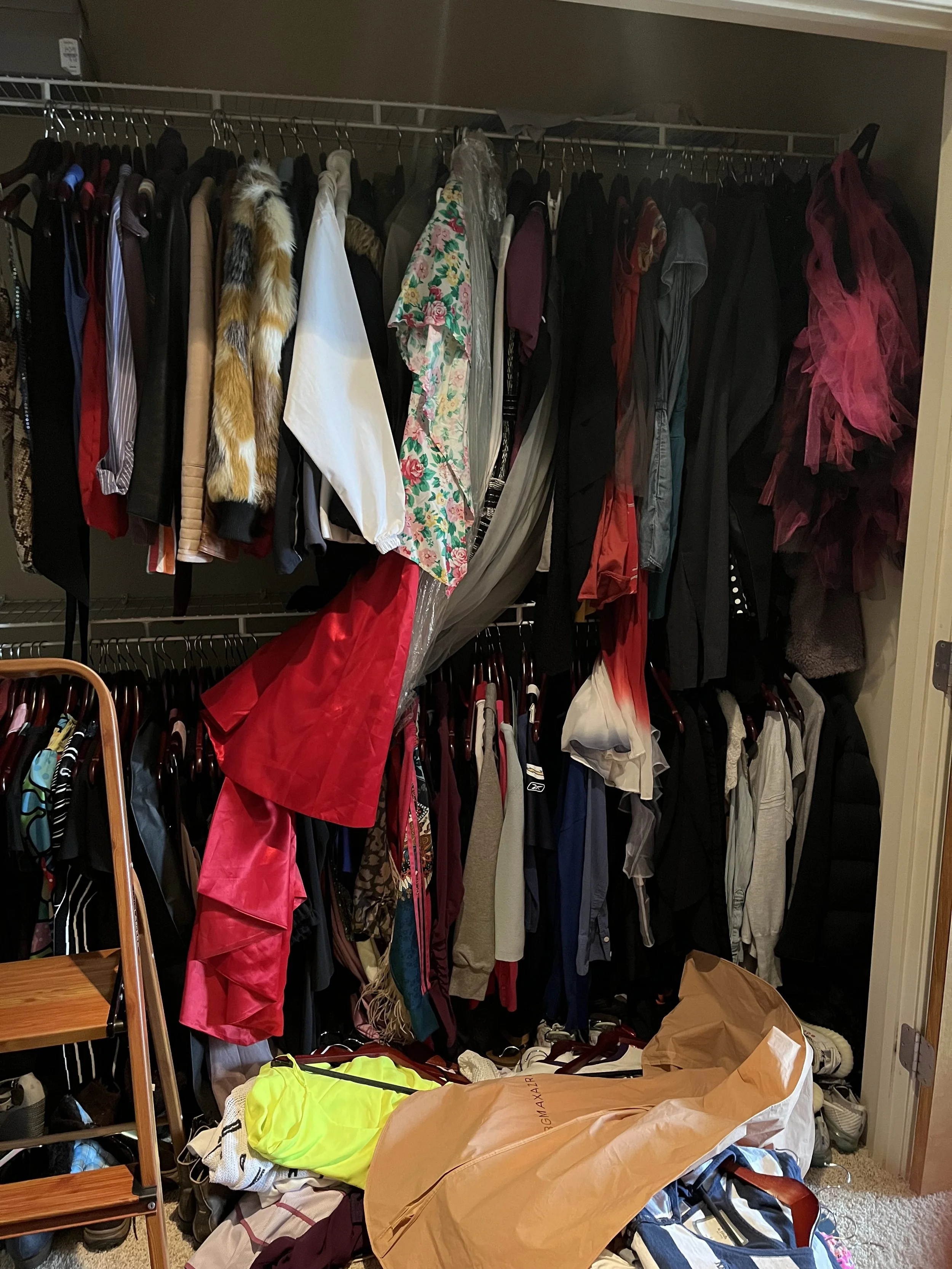 Picture of a closet with a small stepladder to the left with a lower and upper hanging bar with lots of clothes messy and on the floor