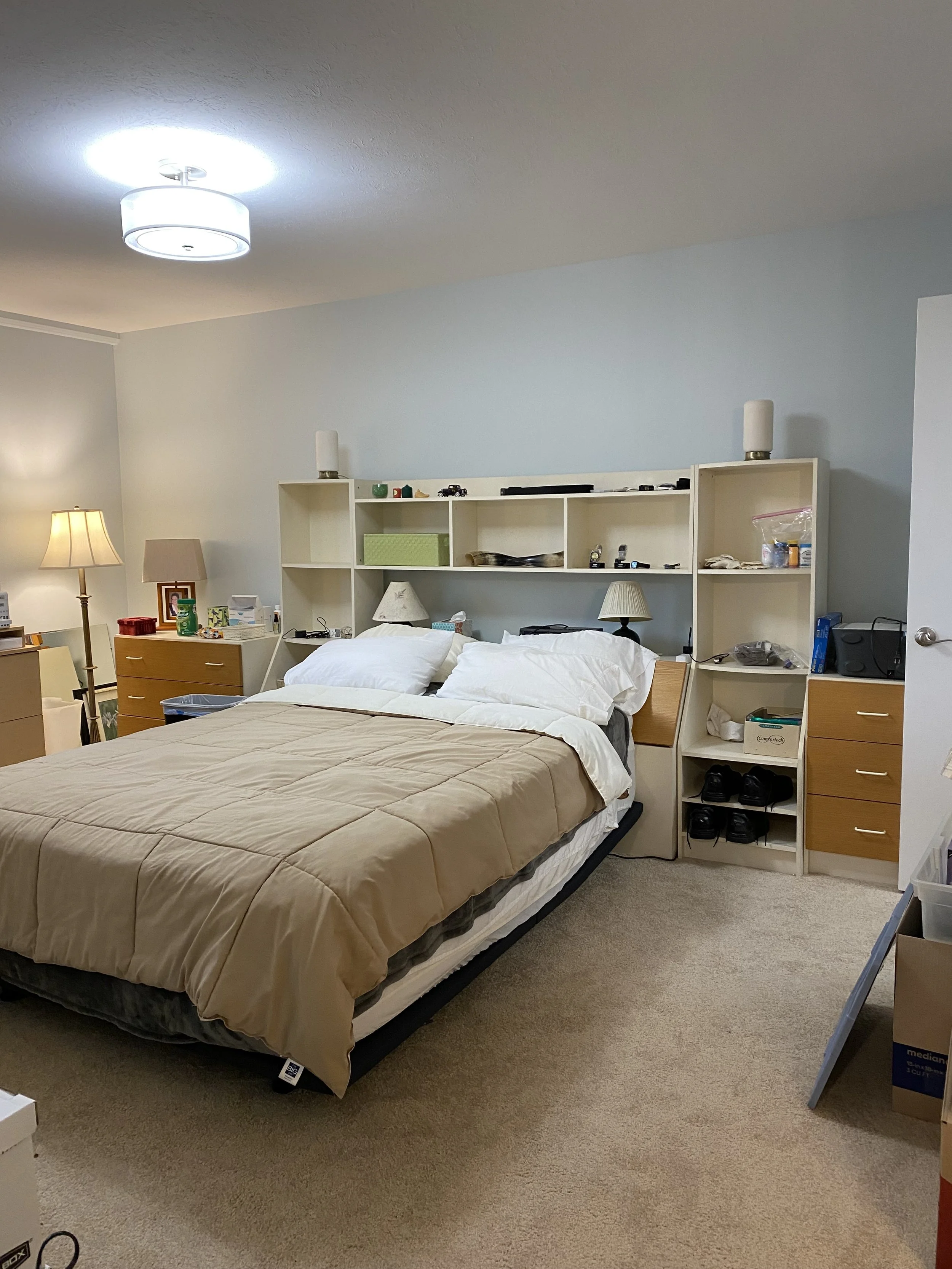 Bedroom with white ceiling light, light blue walls, light brown comforter on a double bed.  Around the bed headboard area is white cubby storage shelves holding lots of small items, small brown night stands drawers at the side