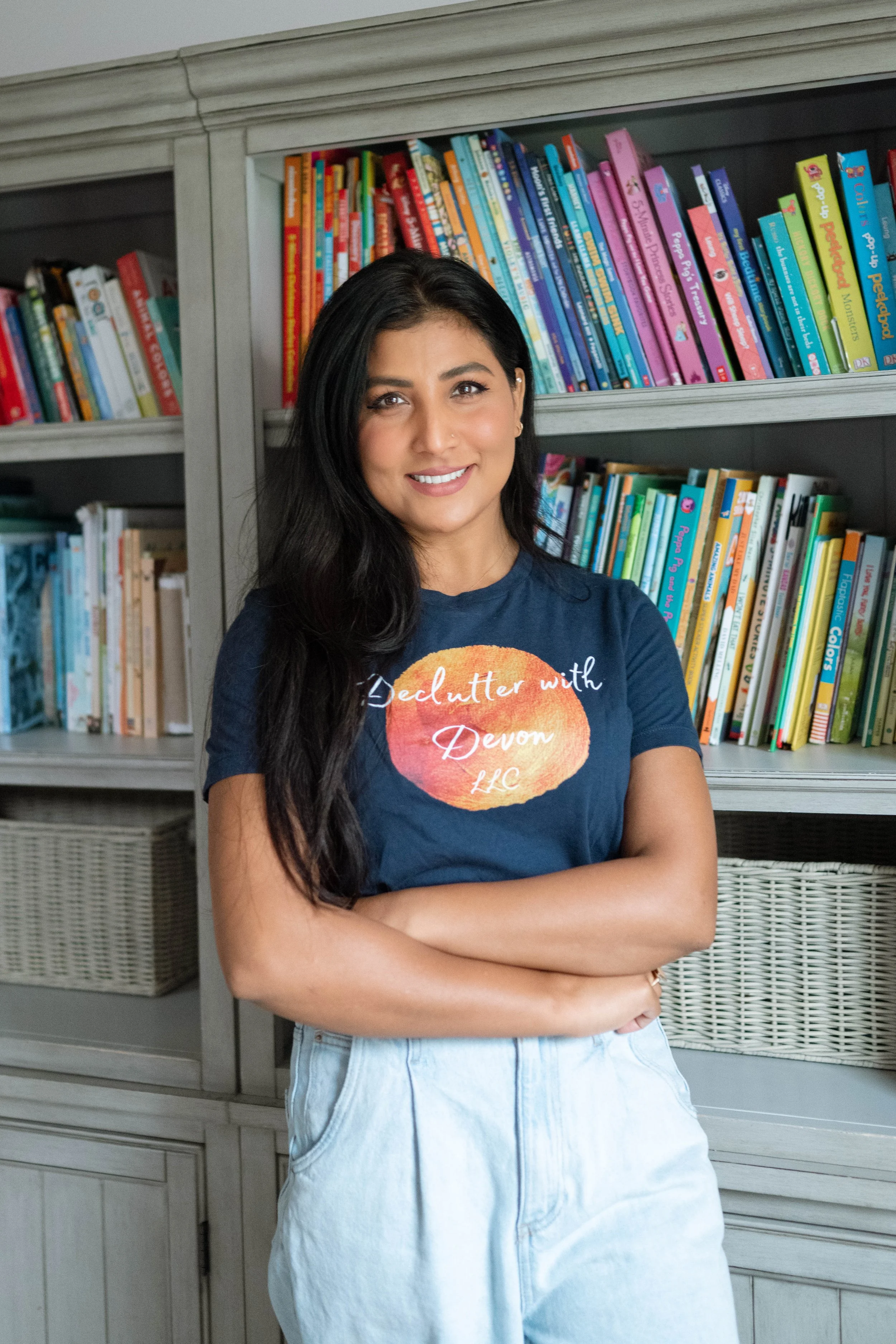 A woman with long black hair, wearing a navy blue t-shirt and light blue jeans, standing in front of a bookshelf filled with colorful children's books, smiling with arms crossed.