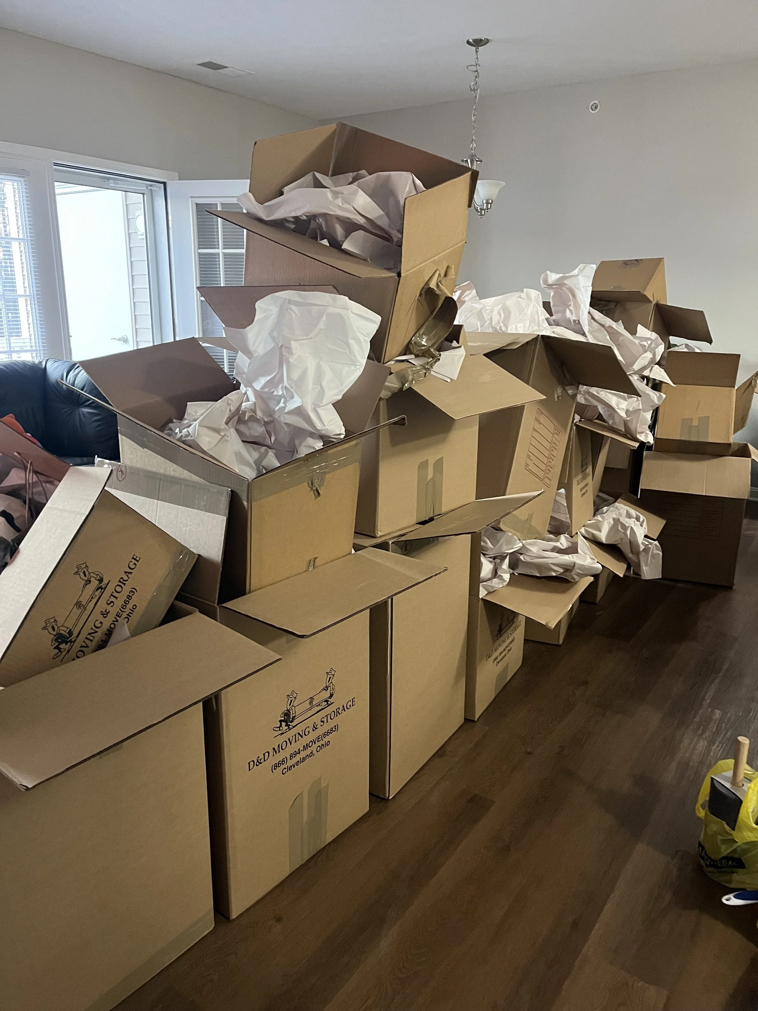 Picture of many large moving boxes with packing paper stuffed and coming out of them on a dark wooden floor with windows seen beyond the large pile.