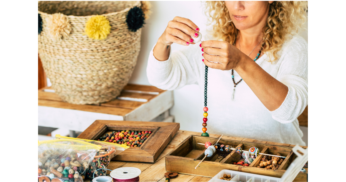 white woman in a white shirt with curly hair holding up a strand of beads with many additional beading supplies on the tables as well as a large woven basket in the background