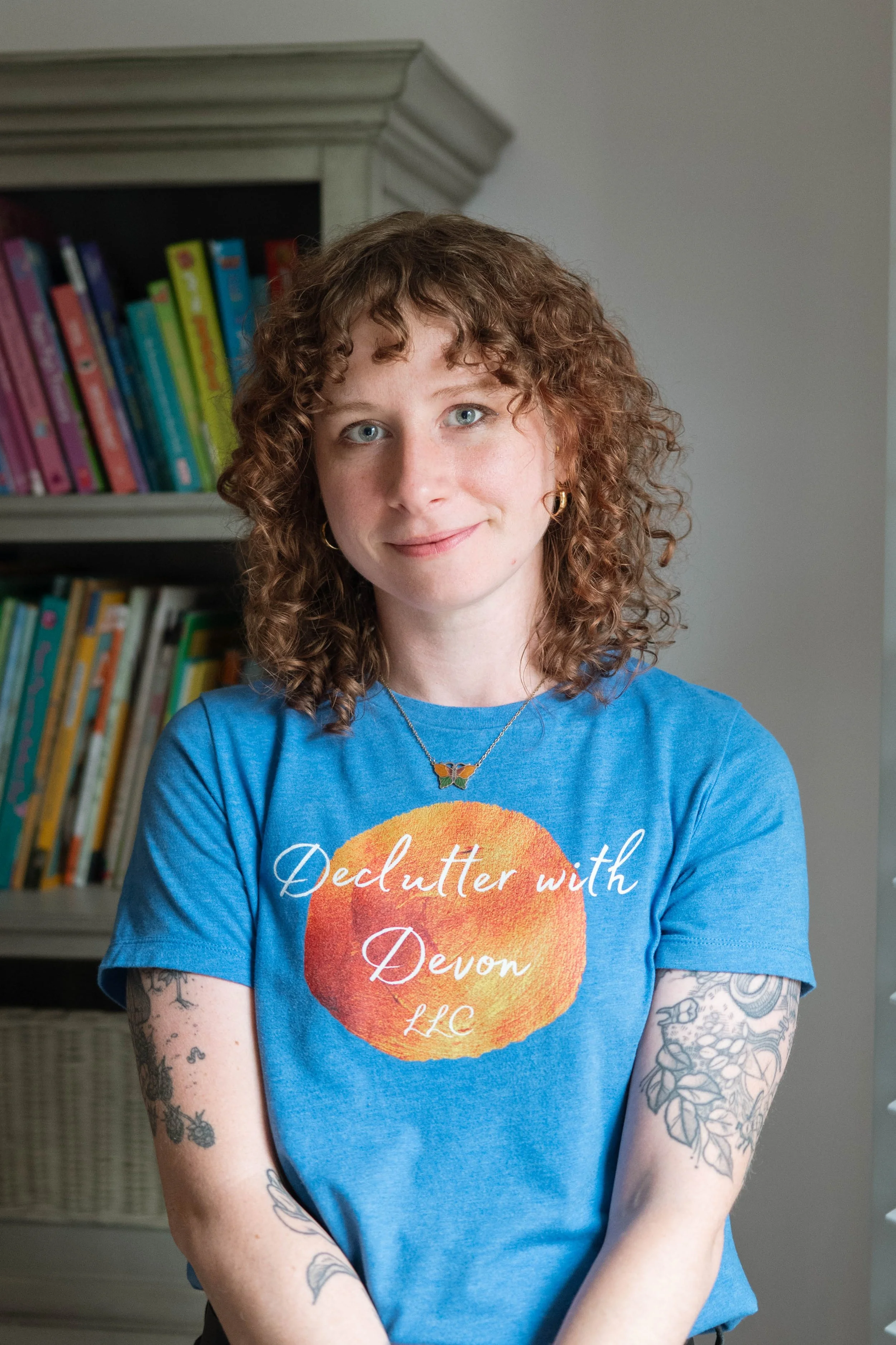 A woman with curly red hair and tattoos on her arms, wearing a blue T-shirt with the text "Declutter with Devon LLC" and a butterfly necklace, standing in front of a bookshelf with colorful books.