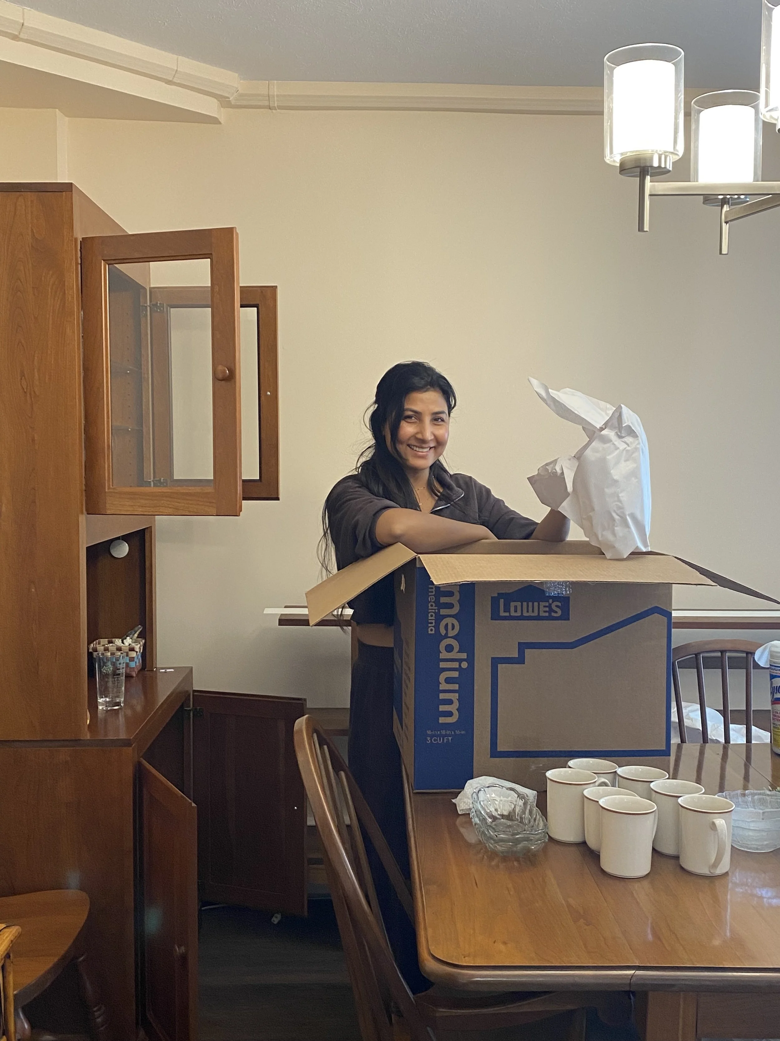 Smiling woman with long dark hair holding paper over a medium sized moving box.  Next to her an open wooden cupboard.  The box is sitting on a dining table with white mugs sitting on the table.