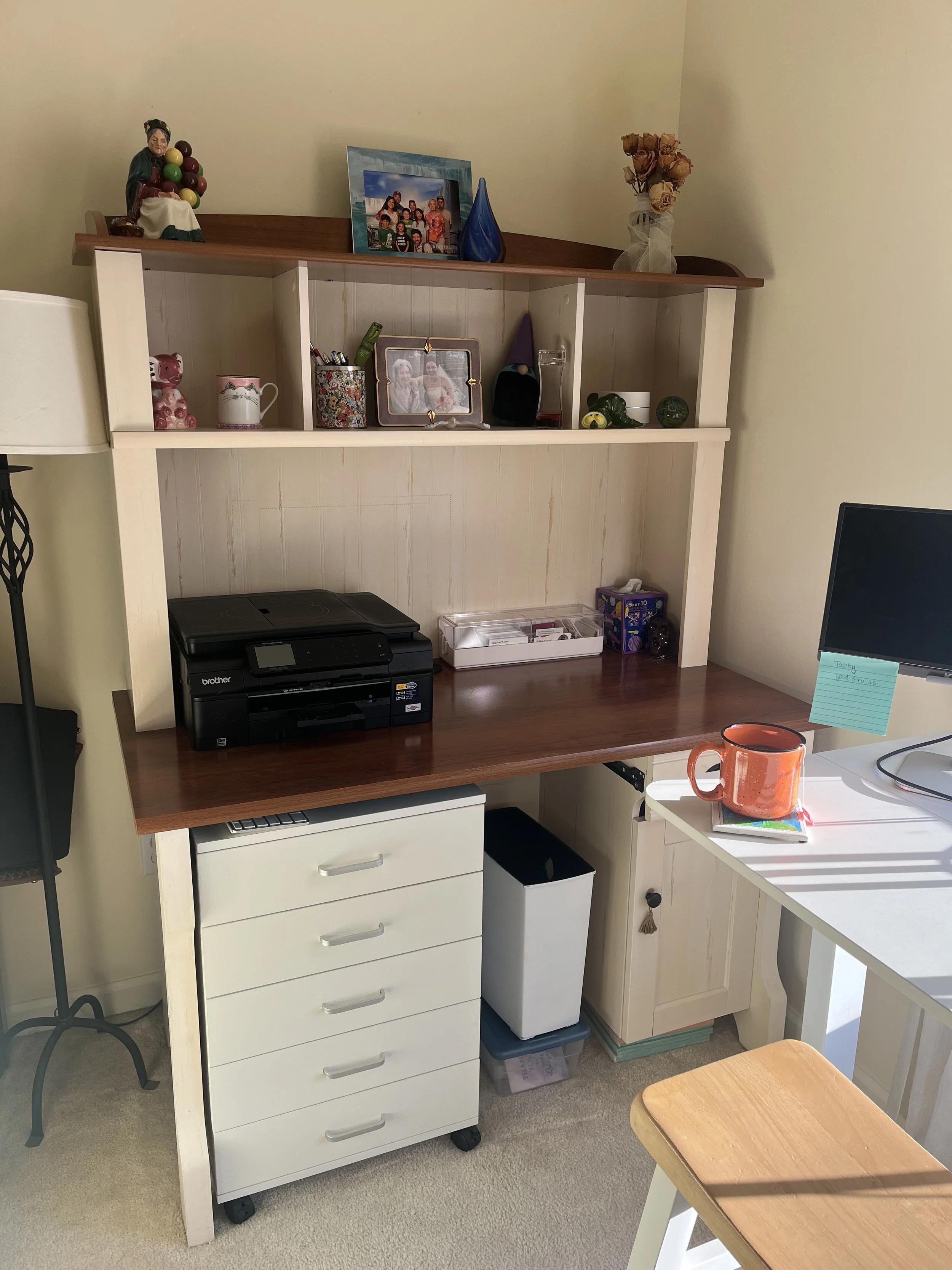 A tidy and organized desk with hutch on it with a set of drawers tucked under and small trash can under and a few tasteful pictures on top.  There's a large black printer sitting on the desk