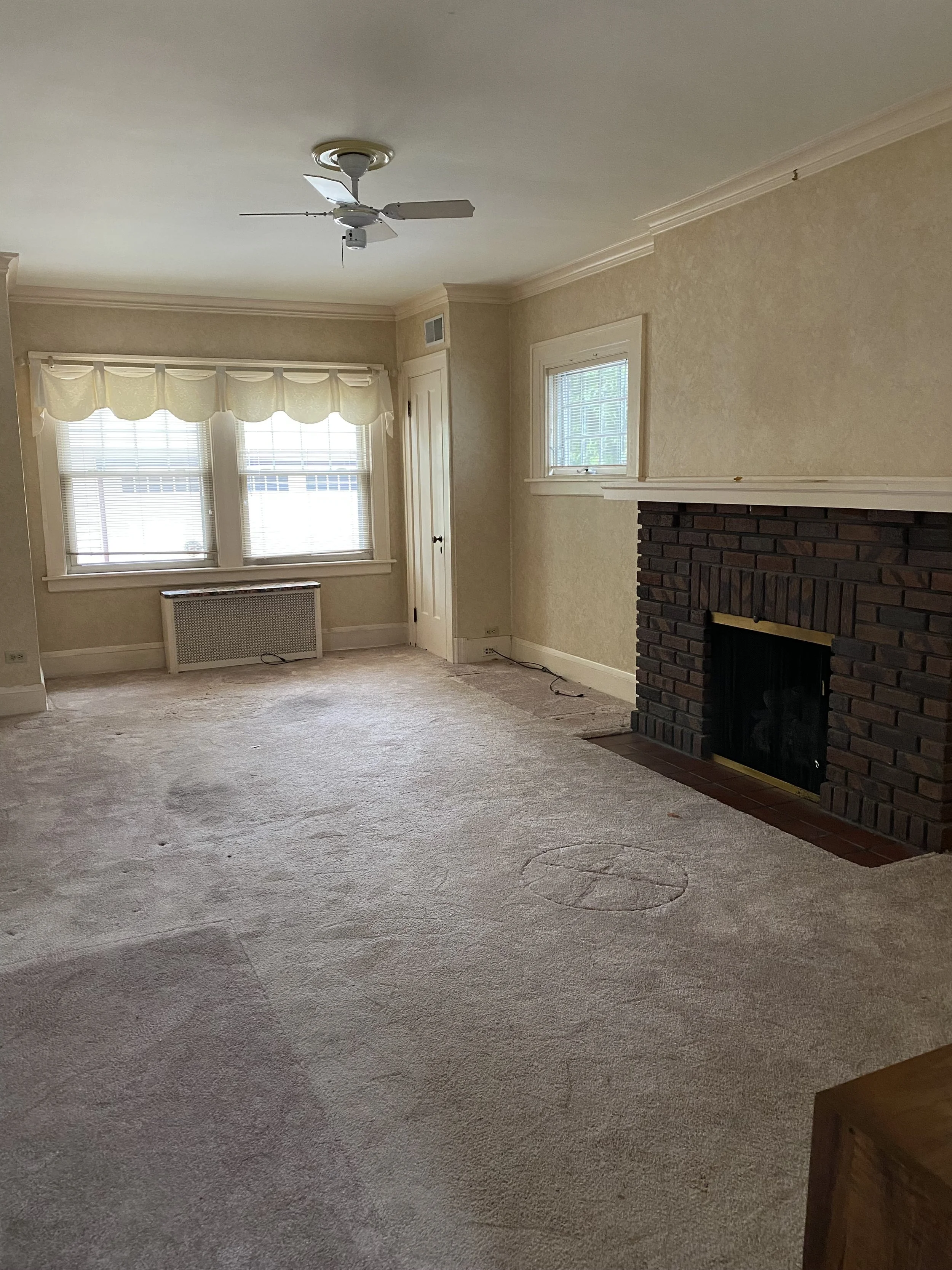 Empty living room before moving without furniture showing a very pale brown carpet and cream walls.  You can see windows at the back of the room over a radiator and cream curtains.  There is a small closet door and a brick fireplace.