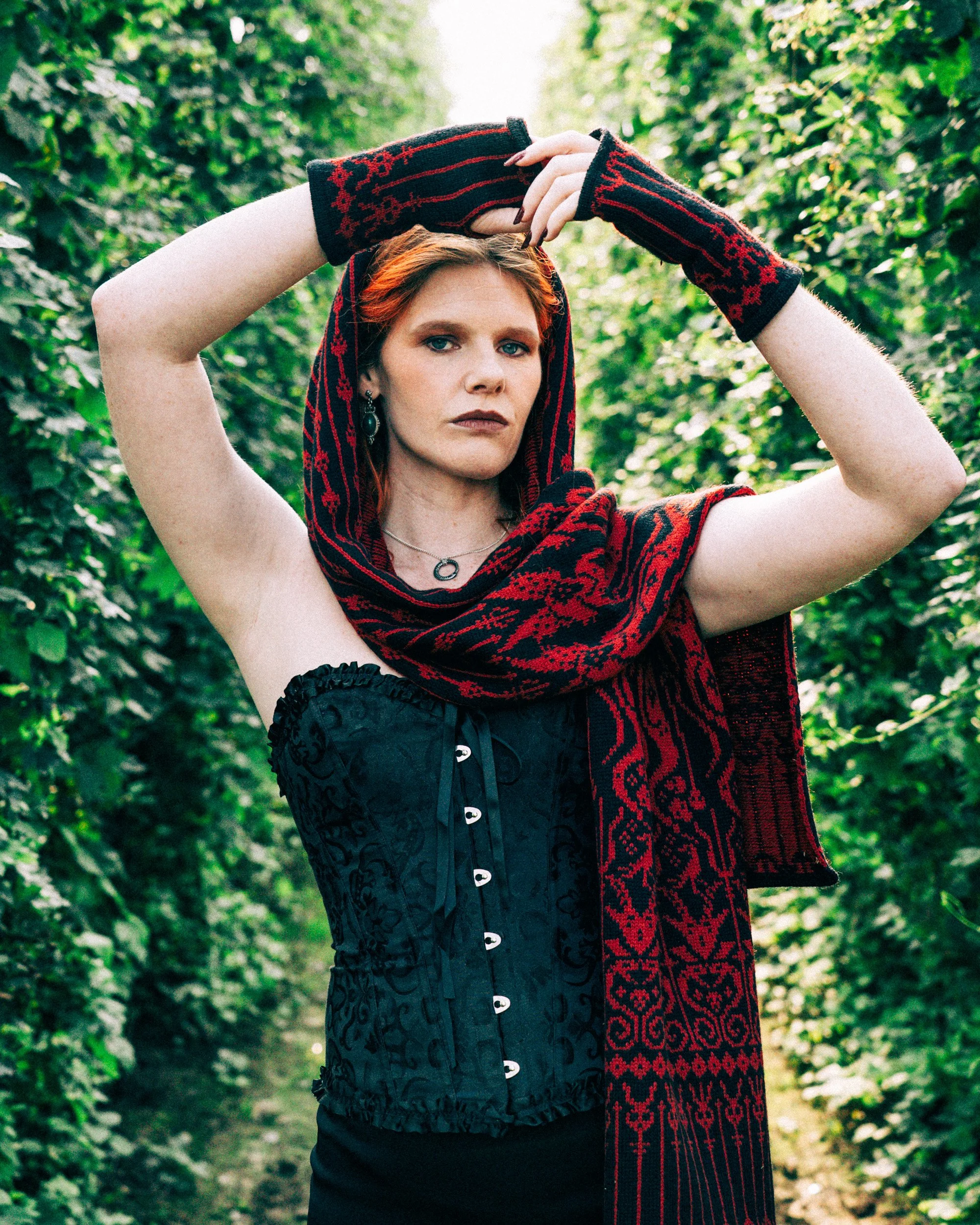 Model wearing Melusine merino wool mittens in black and red, designed to coordinate with the Melusine scarf, photographed in a woodland garden.