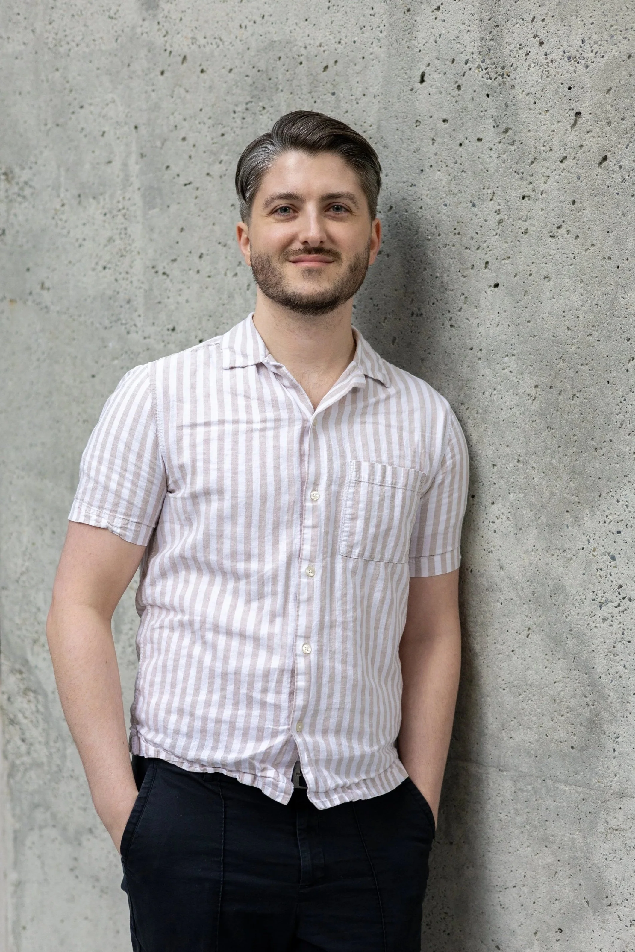 Jacob Stinson, a young man with gray hair and a beard stands against a textured concrete wall, wearing a short-sleeved striped shirt and dark pants, with his left hand in his pocket and a slight smile on his face.