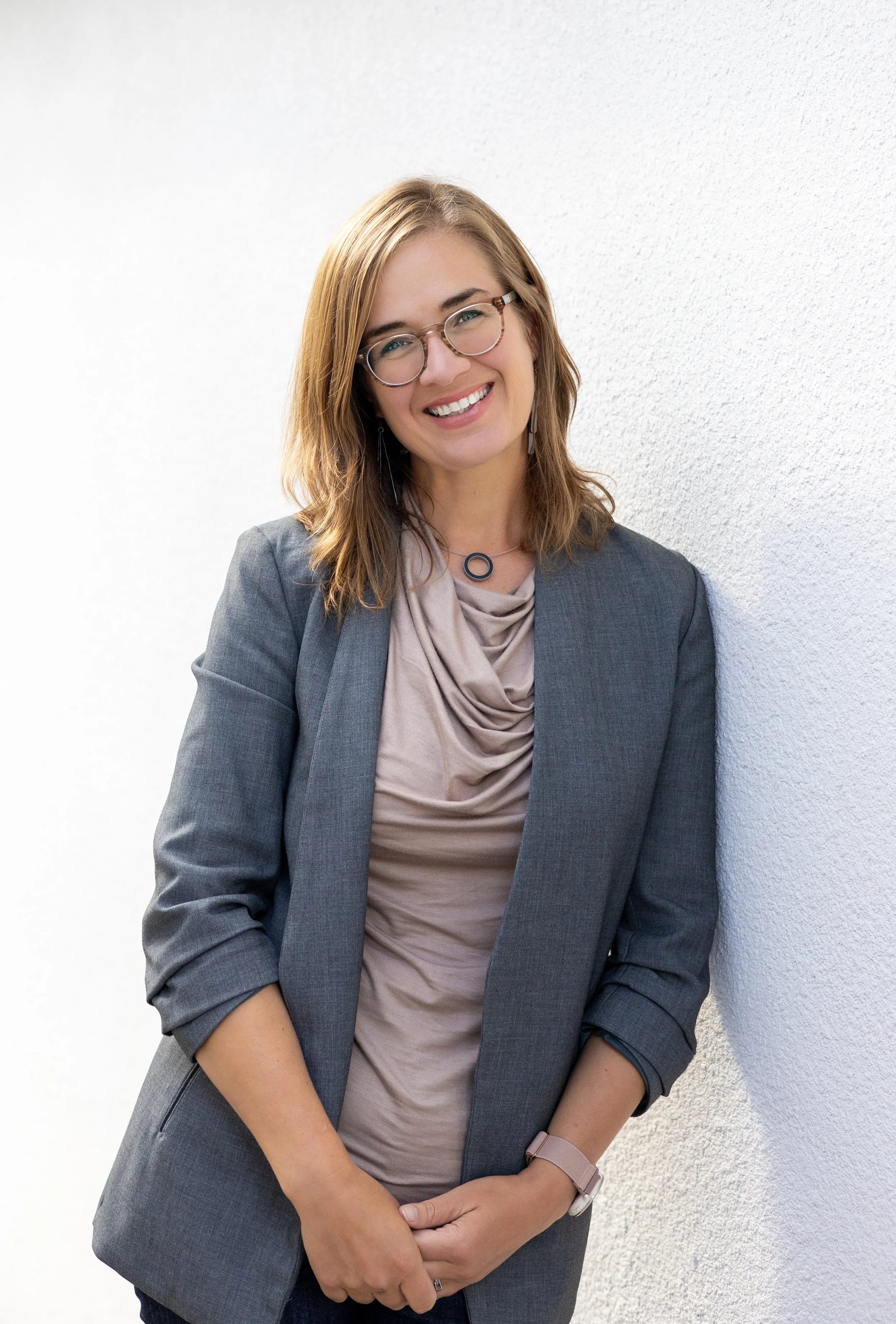 Portrait of Melissa Maddux Renner, a smiling woman with glasses, wearing a gray blazer and beige blouse, standing against a white textured wall.