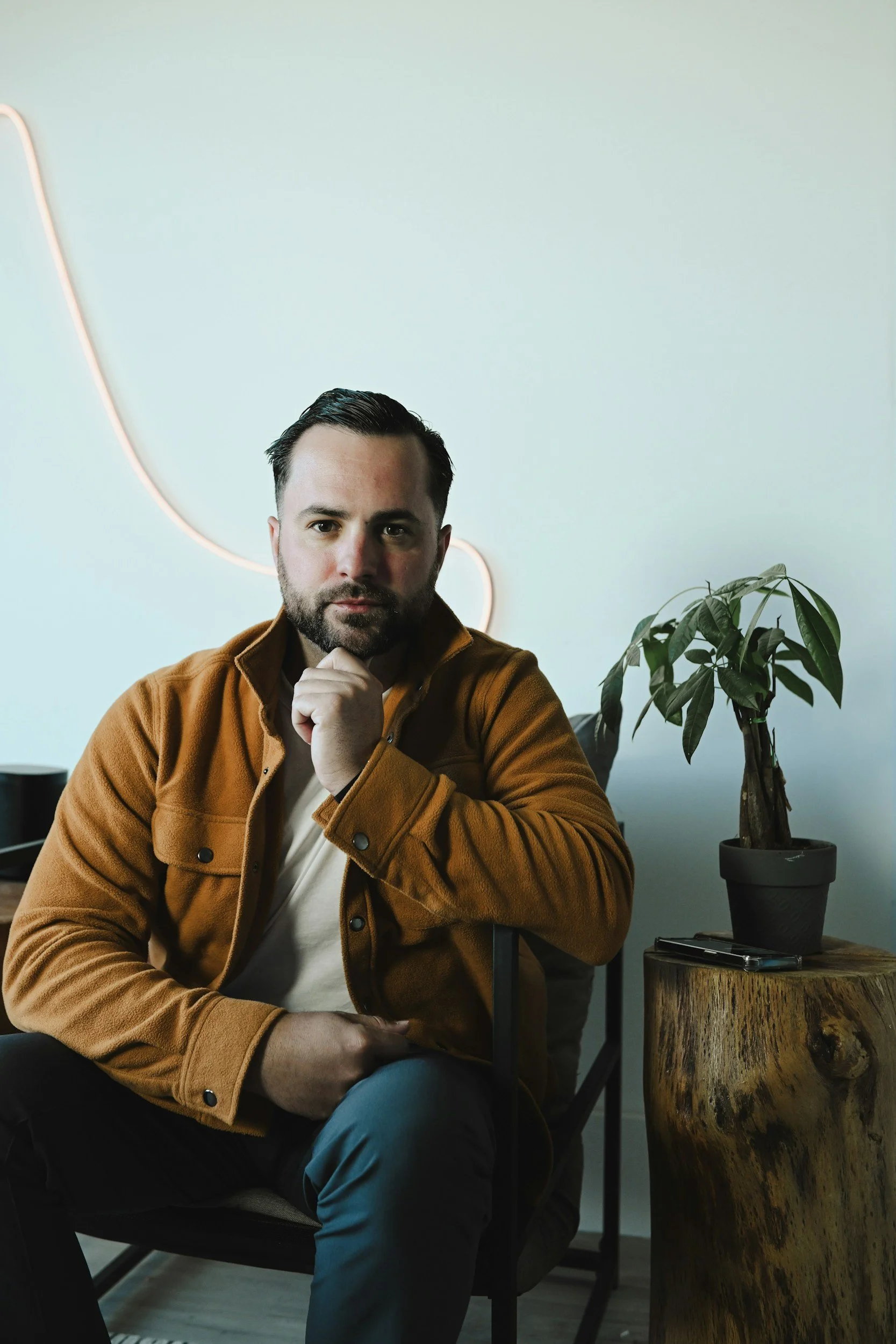 A man with dark hair and beard wearing a brown jacket sitting on a chair next to a wooden side table with a potted plant on it. He is looking directly at the camera with his hand resting on his chin.