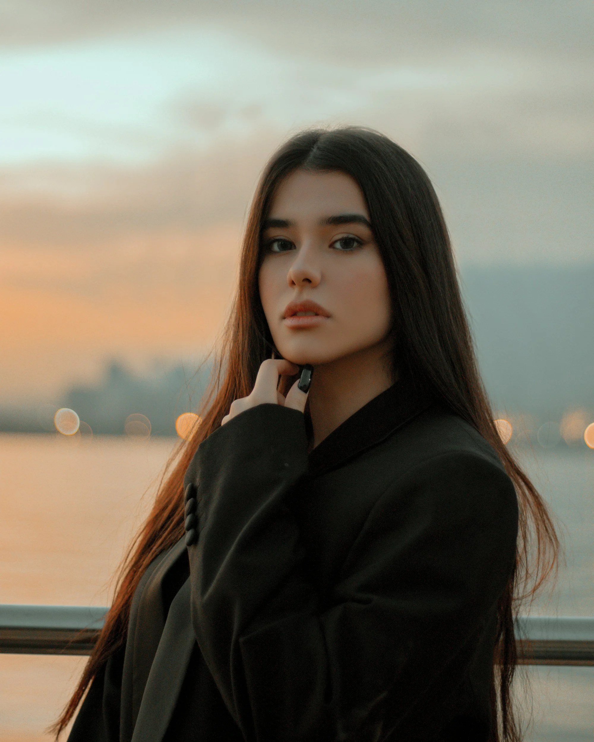 A young woman with long dark hair and fair skin posing outdoors during sunset, wearing a black blazer, with a water body and cityscape in the background.