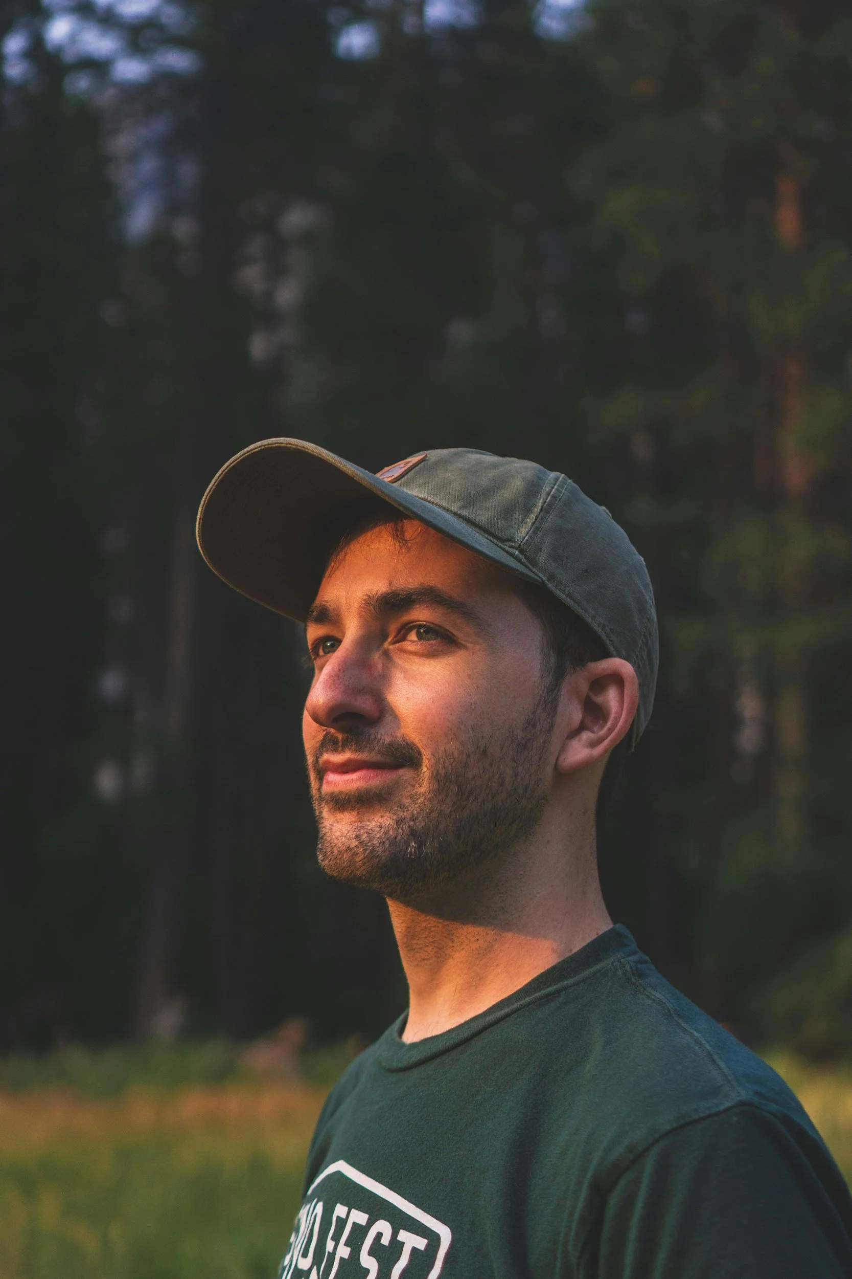 A man with a beard wearing a dark green T-shirt and a baseball cap outdoors during sunset, with a background of trees.