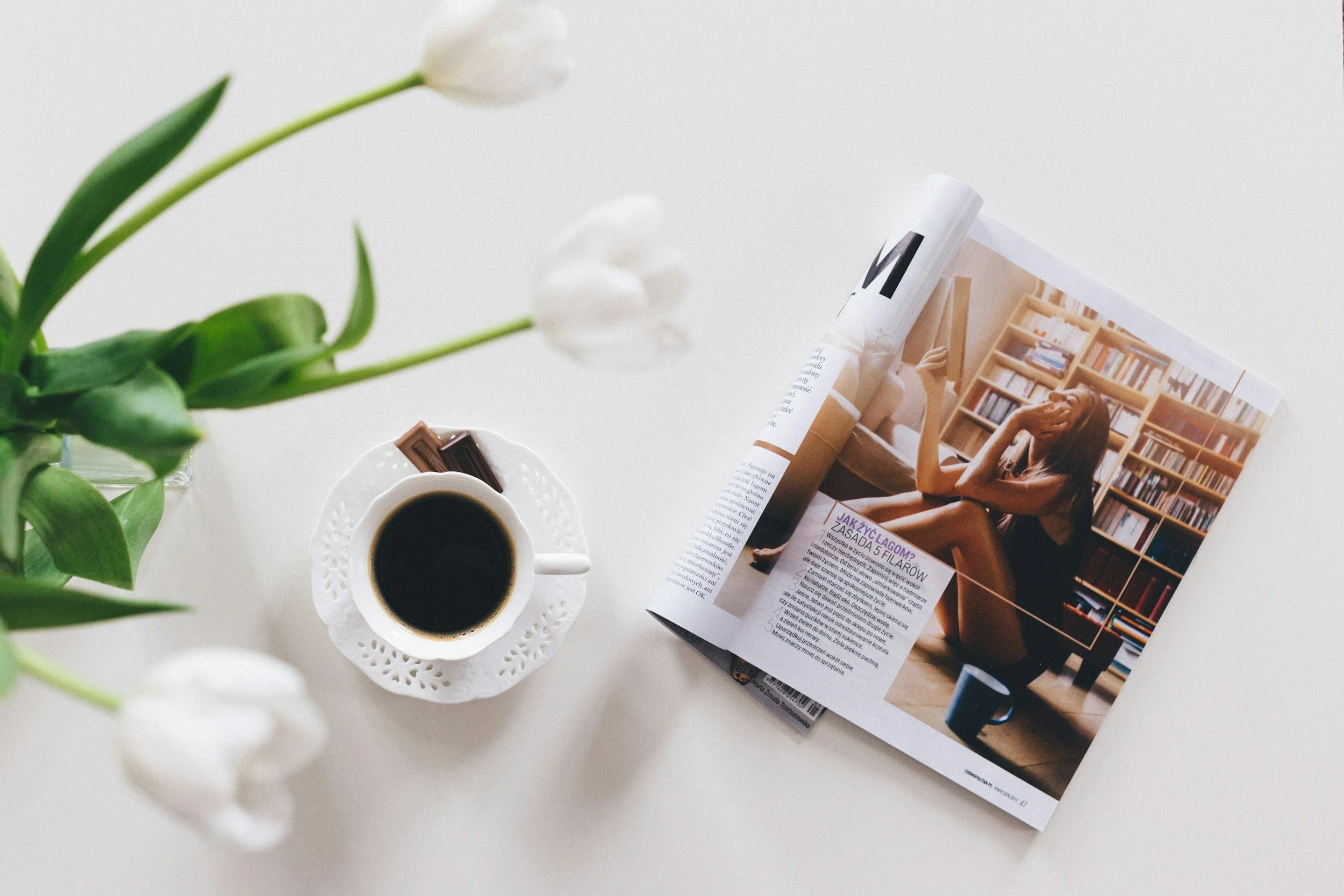 A white table with a vase of white tulips, a cup of black coffee on a decorative saucer, and an open magazine showing a woman sitting by a bookshelf.