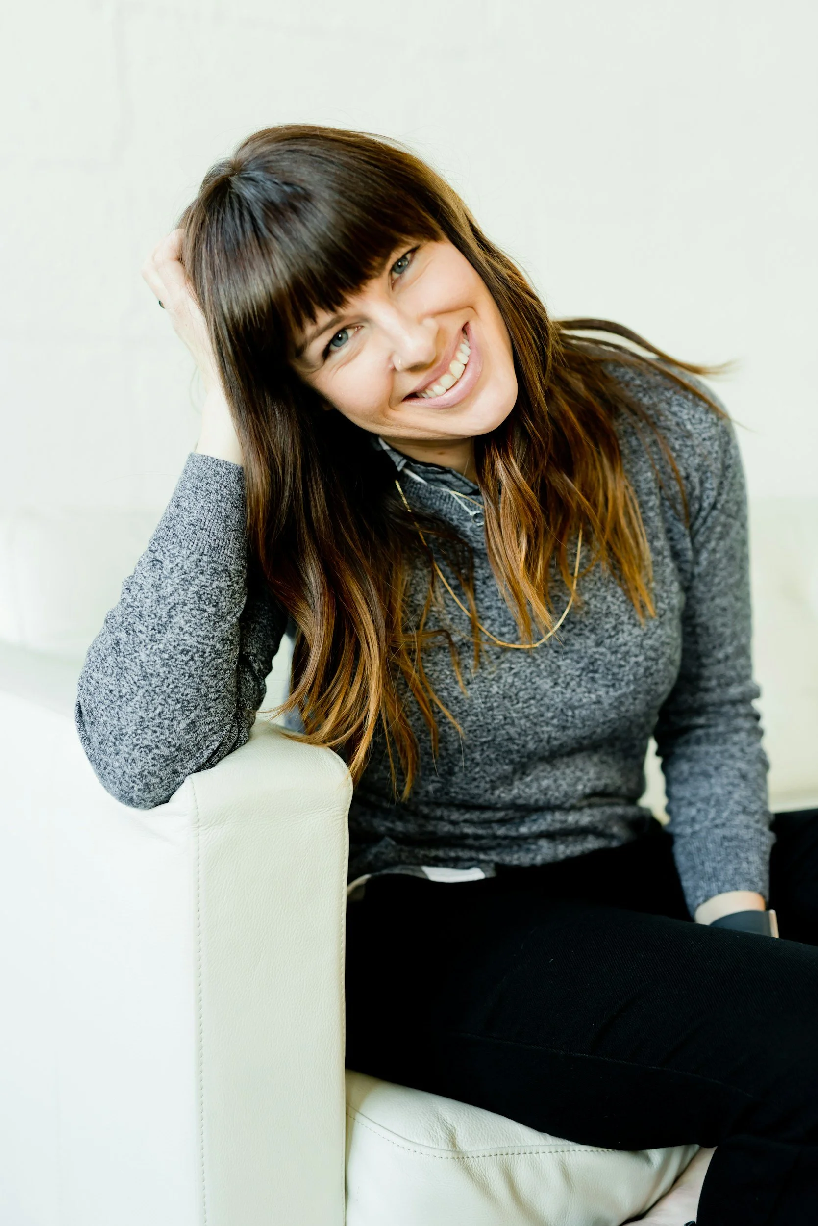 A woman with long brown hair and bangs, smiling and leaning on a white sofa, wearing a gray sweater and black pants.