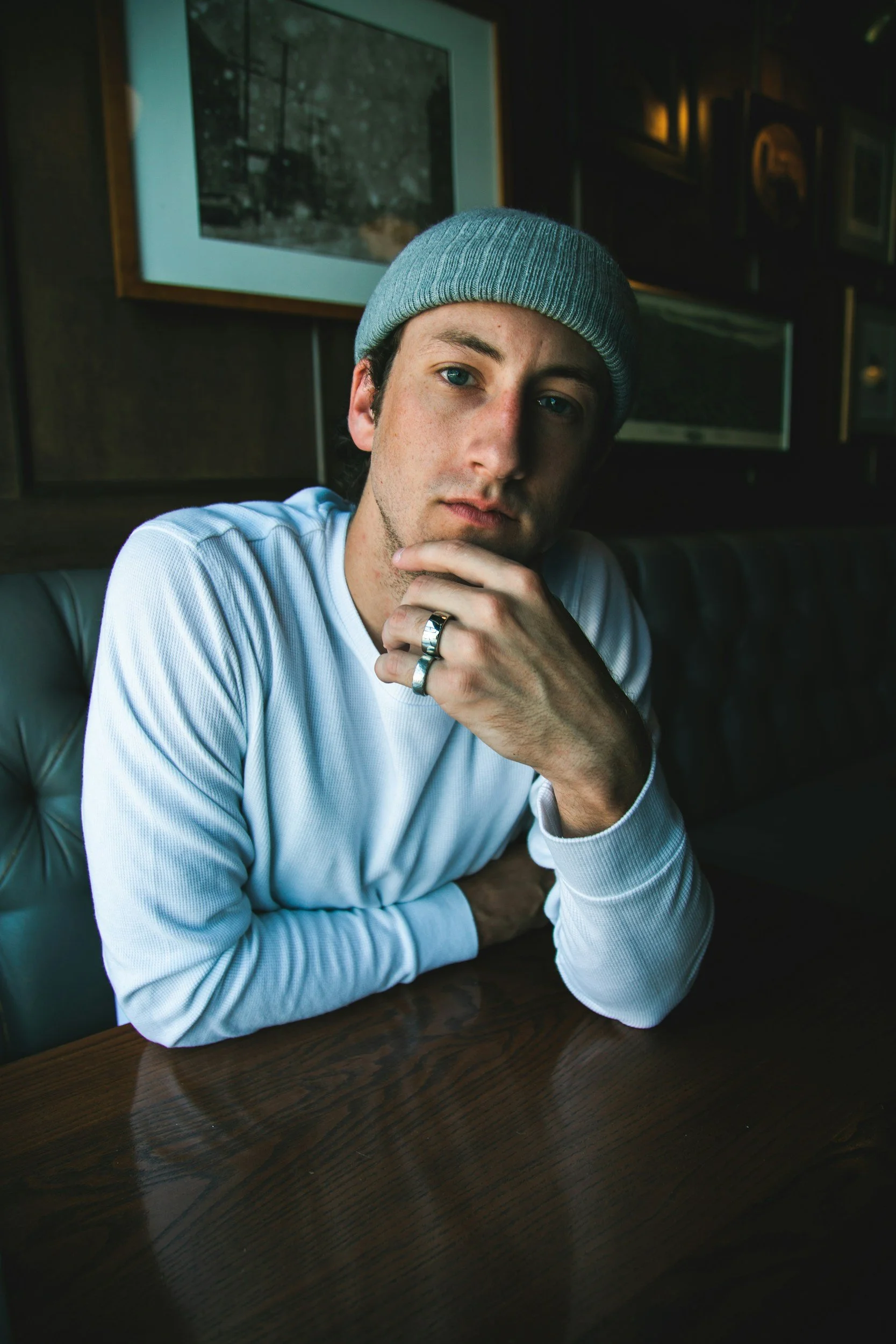 A young man wearing a gray beanie and a white long-sleeve shirt sits at a wooden table in a dimly lit room, resting his chin on his hand and looking directly at the camera.