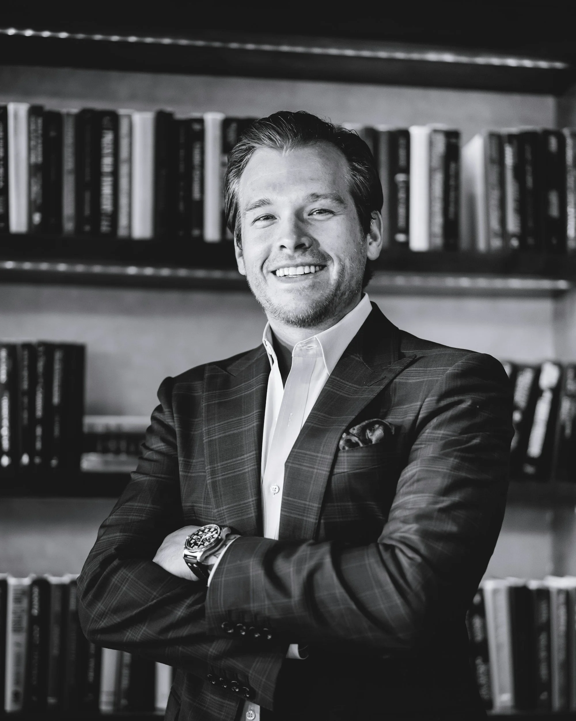 A professional man with light hair and a beard, dressed in a checkered suit jacket and white shirt, smiling with arms crossed in front of a bookshelf.