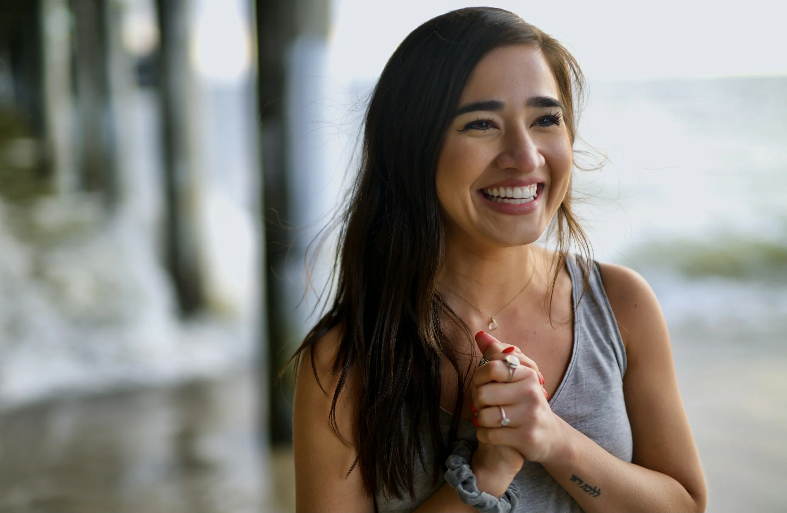 A young woman with long dark hair, smiling with her hands clasped together, standing on a beach or pier with the ocean and wooden posts in the background.