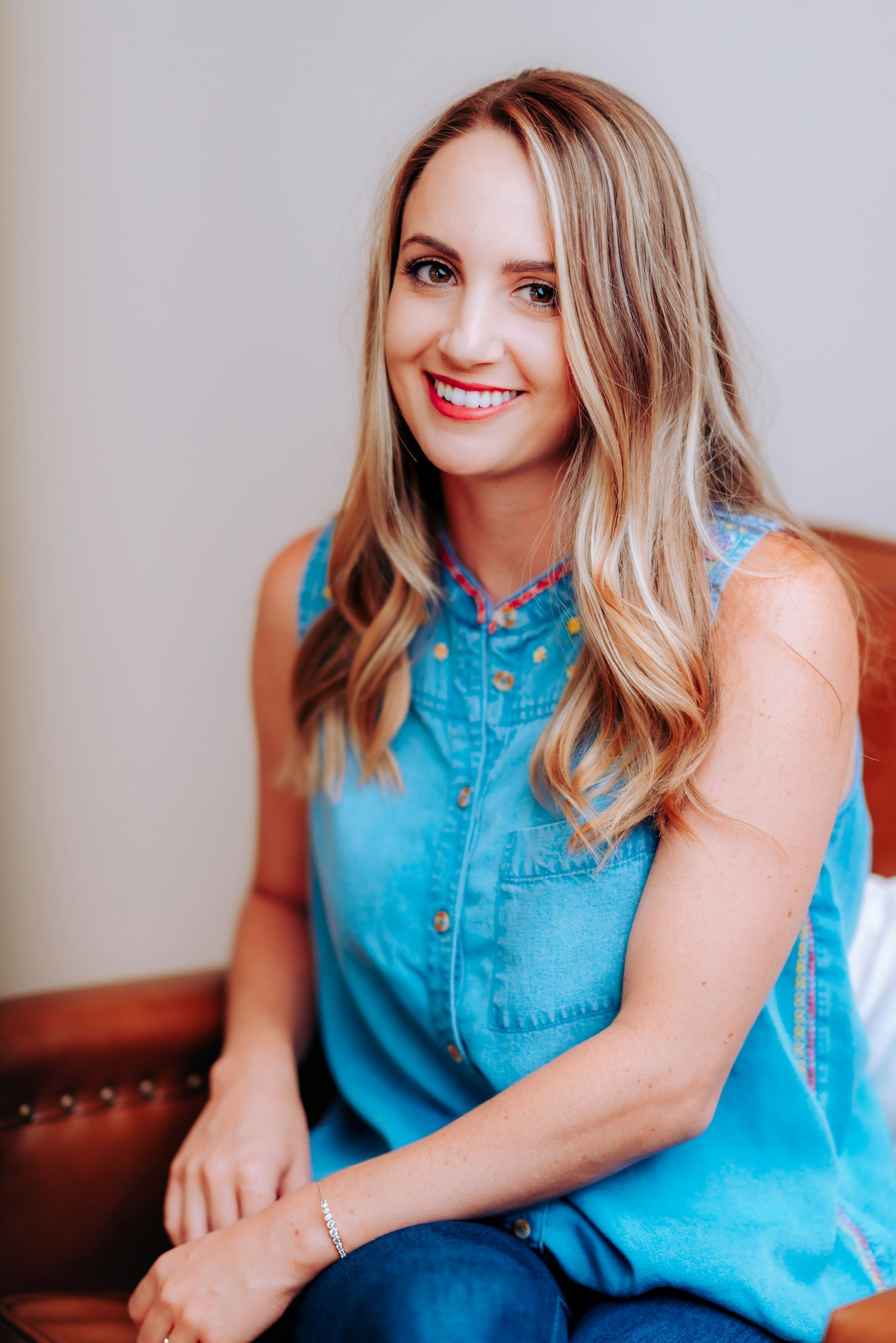 A woman with light brown hair styled in loose waves, wearing a sleeveless blue denim top, sitting on a brown leather couch and smiling at the camera.