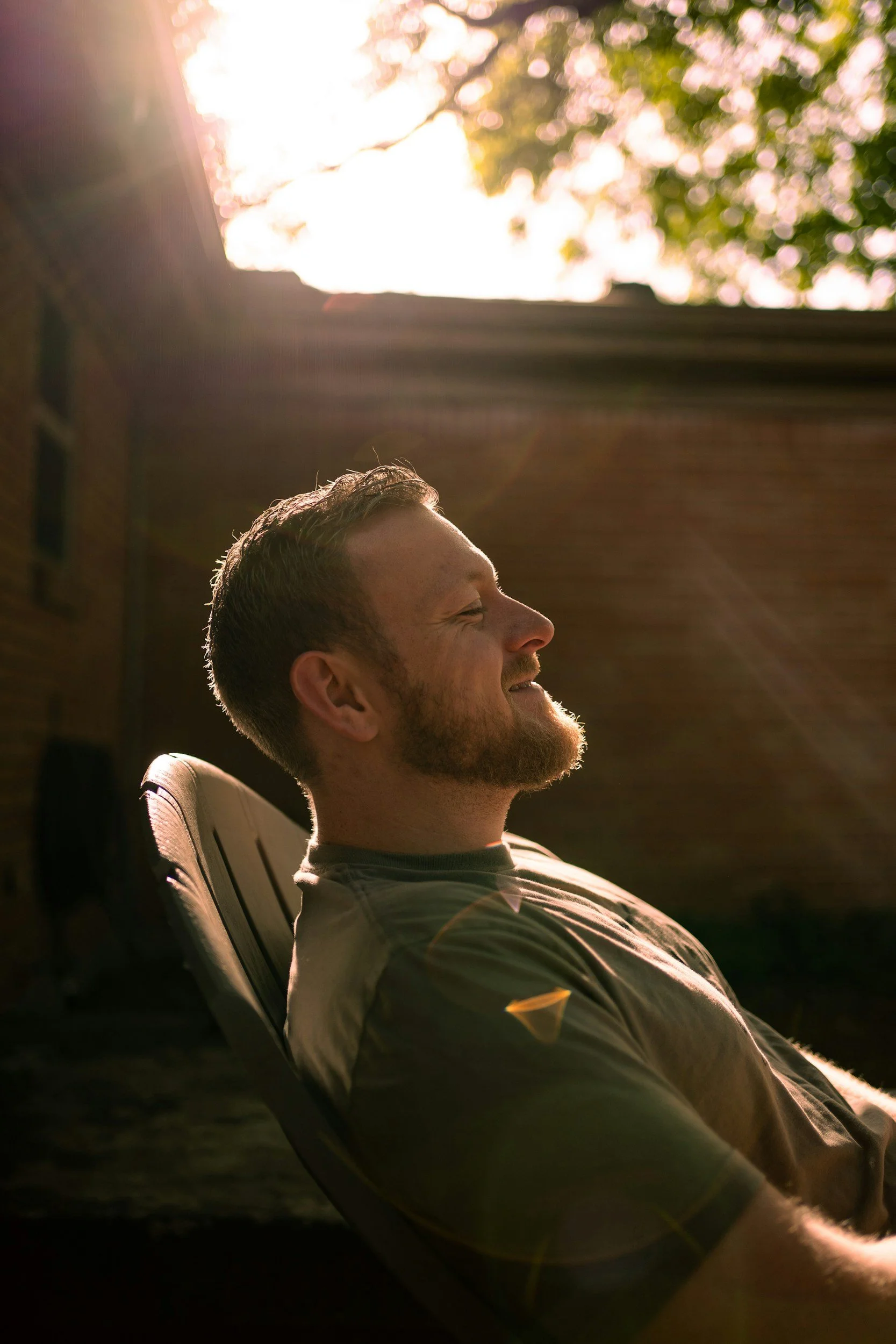 A man with a beard and short hair relaxing in a chair outdoors during sunset, with trees and bright sunlight in the background.