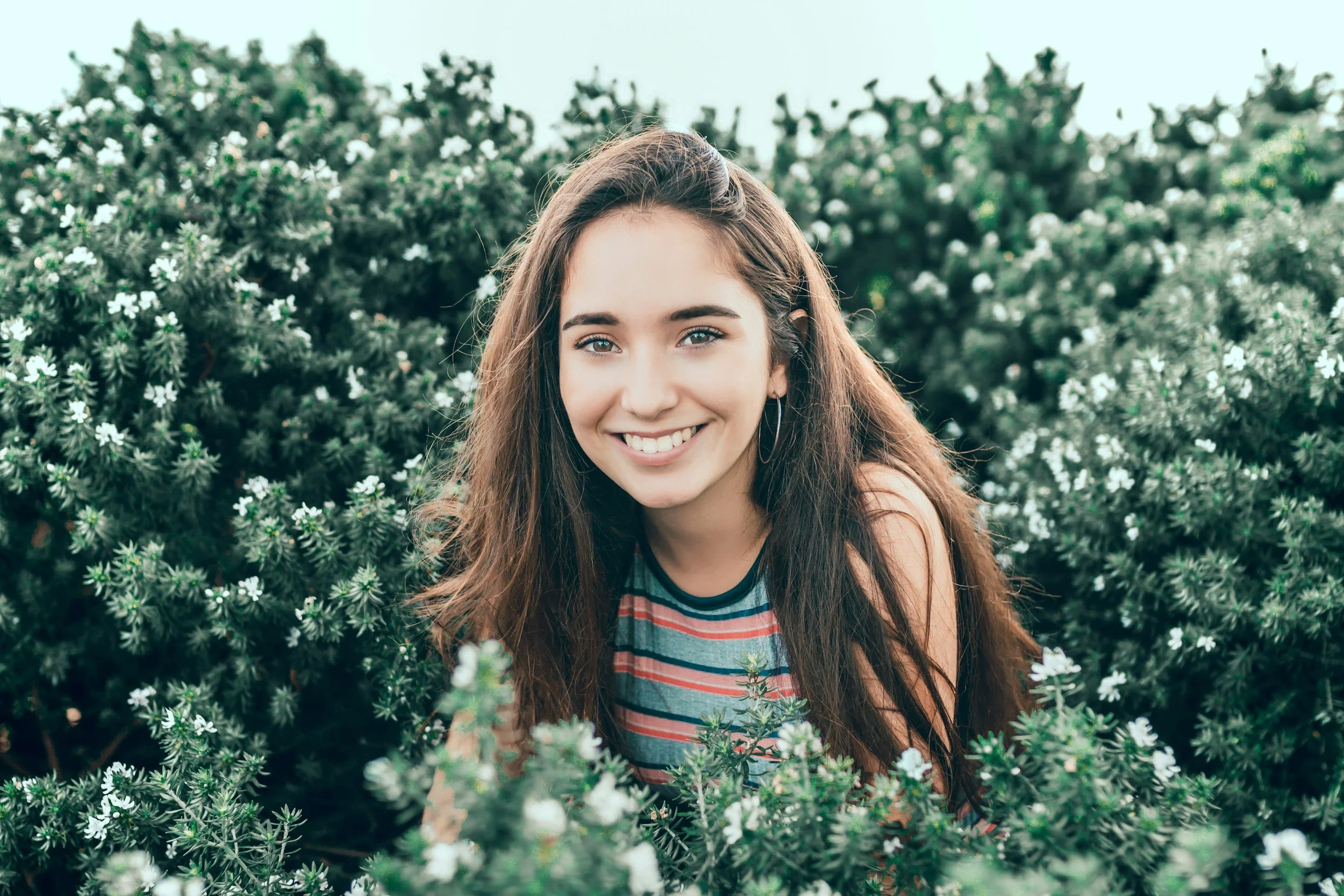 A young woman with long brown hair, smiling, is standing outdoors among green bushes with small white flowers, wearing a striped t-shirt.