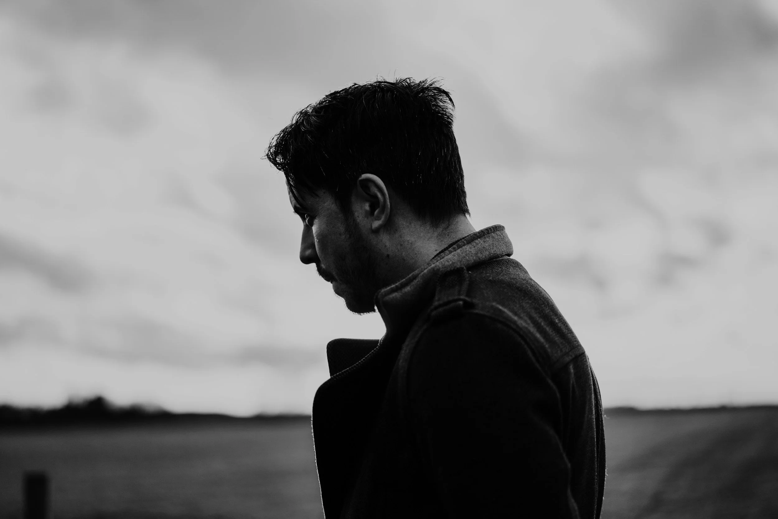 A black and white profile of a man with dark hair wearing a jacket, looking down or away, outdoors with a cloudy sky in the background.