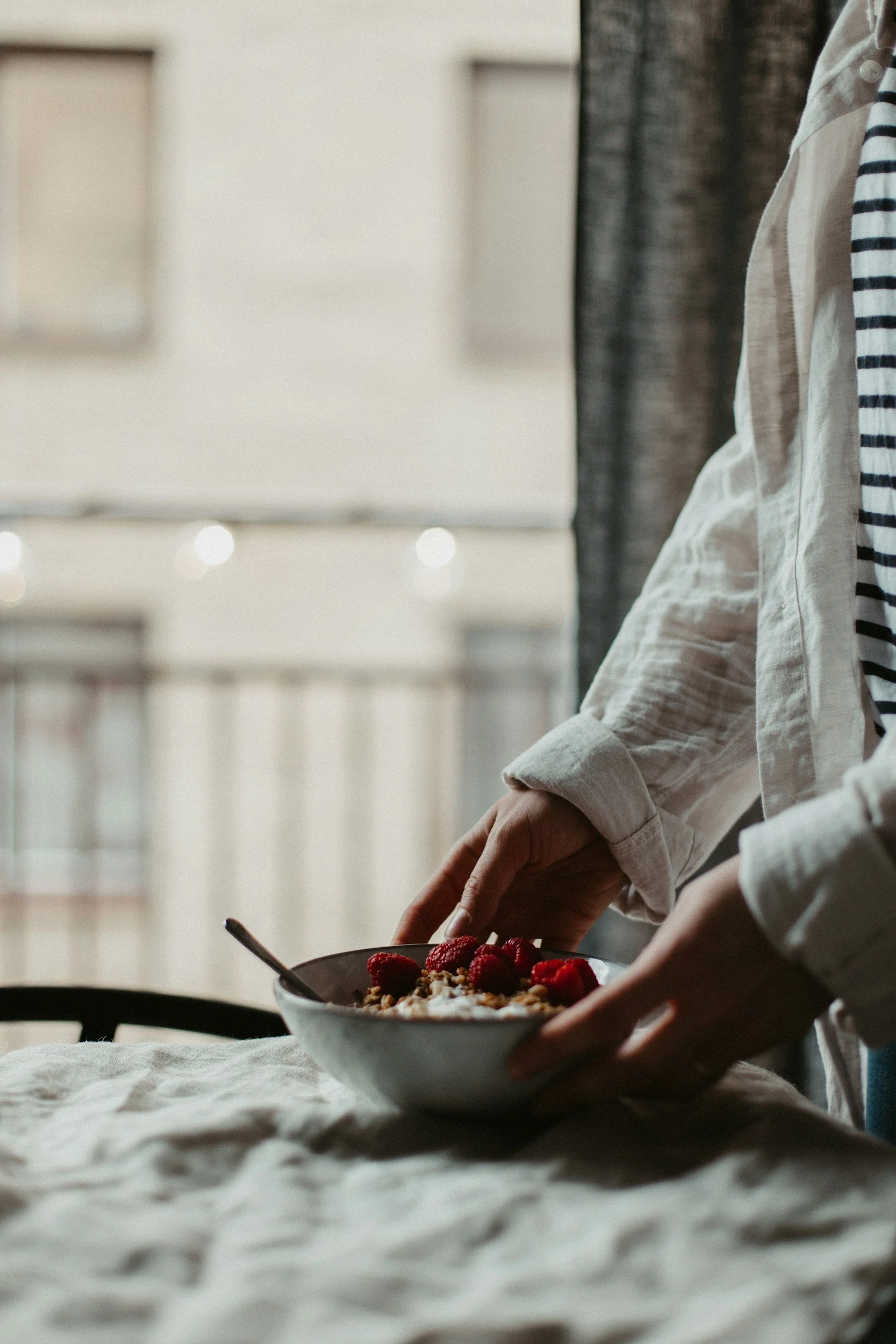 A person in a white shirt with rolled-up sleeves is leaning over a table, holding a bowl of cereal topped with raspberries and other berries, with a spoon inside. The background shows a brightly lit room with large windows.