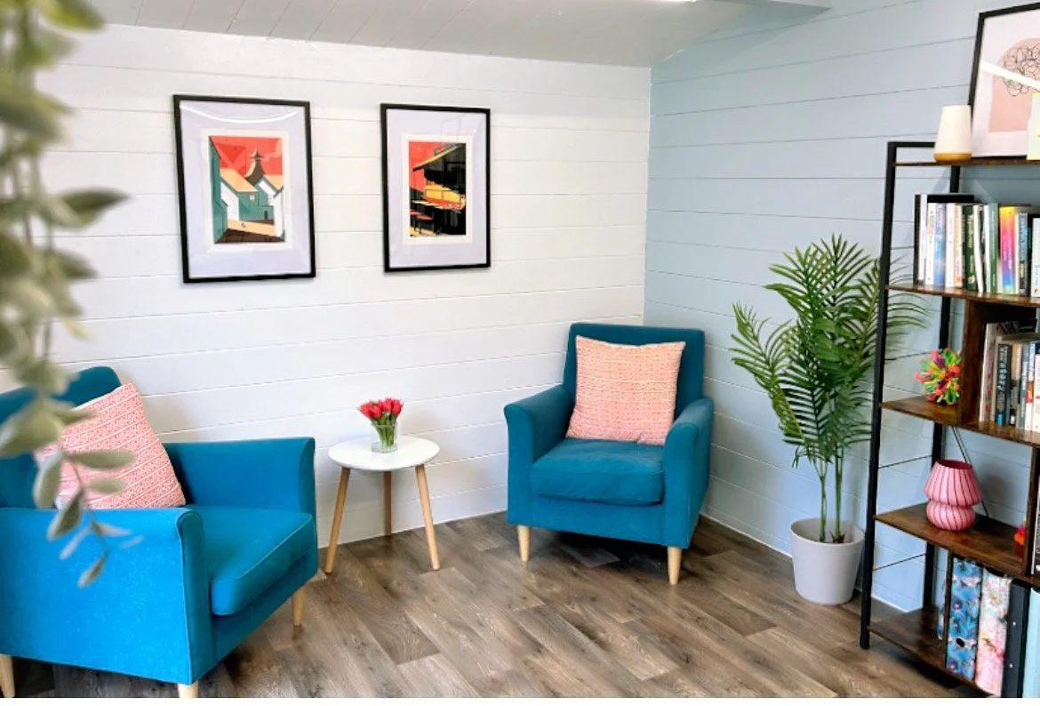 Cozy living room with two blue armchairs, coral patterned pillows, a small white table, a vase of red tulips, framed art, and wood flooring.