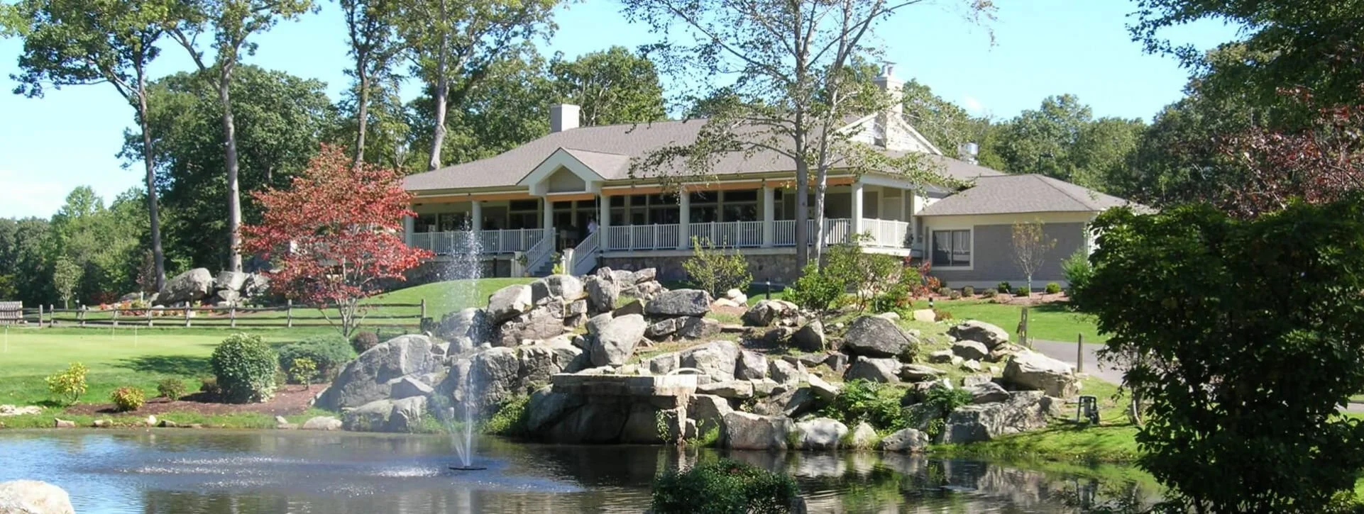 Large house with a wraparound porch overlooking a landscaped yard with rocks, trees, and a pond with a fountain.