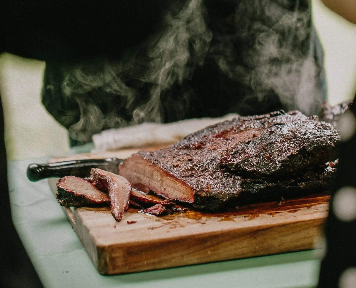A smoked brisket with a smoky crust resting on a wooden cutting board, partially sliced, with steam rising from it.