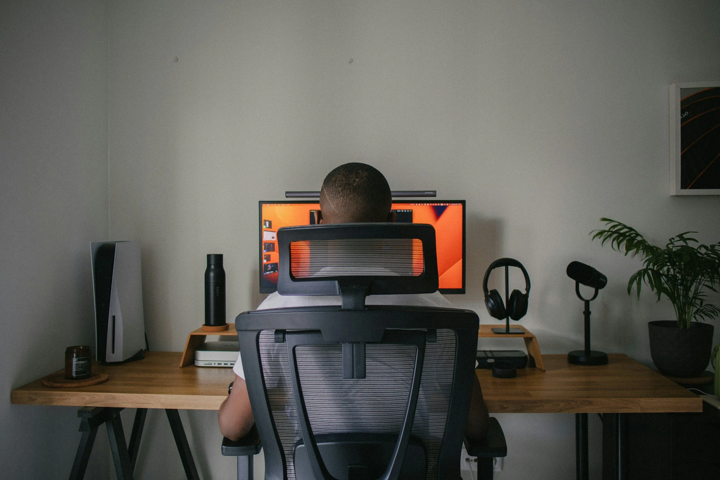 Person working at a wooden desk with a computer monitor, headphones, plant, and various tech accessories in a minimalist home office.