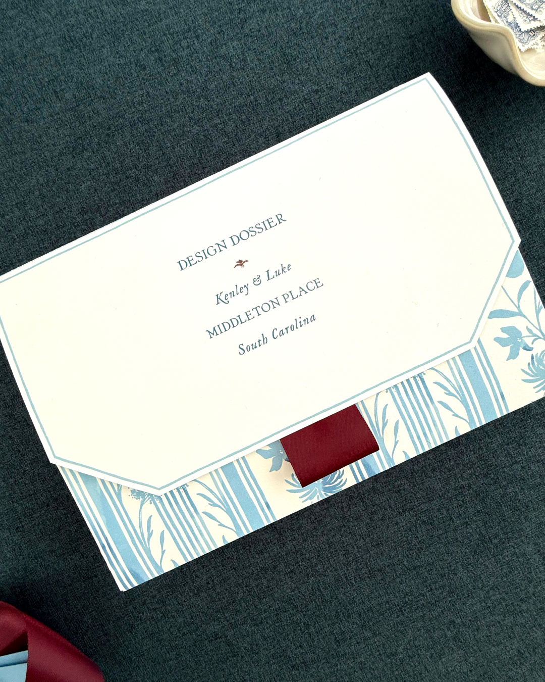 A cream-colored folder labeled “Design Dossier” with blue floral and striped borders and a maroon ribbon accent. The cover includes personalized names and venue details.