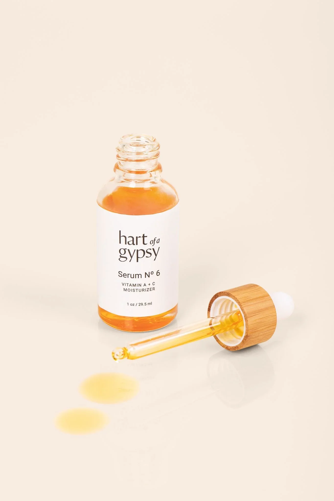 Open amber bottle of “hart of a gypsy Serum No. 6 Vitamin A + C Moisturizer” with a wooden‑topped dropper lying beside it and drops of orange serum on a beige surface.