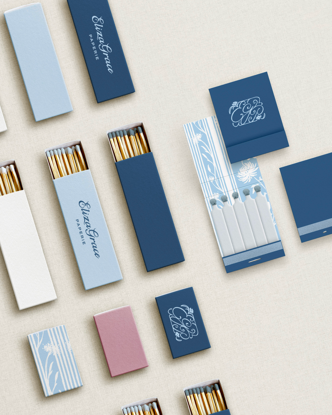 An arrangement of matchboxes and matchbooks in shades of blue, white, and pink, each featuring refined designs and “Eliza Grace Paperie” branding. Some boxes are open, revealing wooden matches with colored tips.