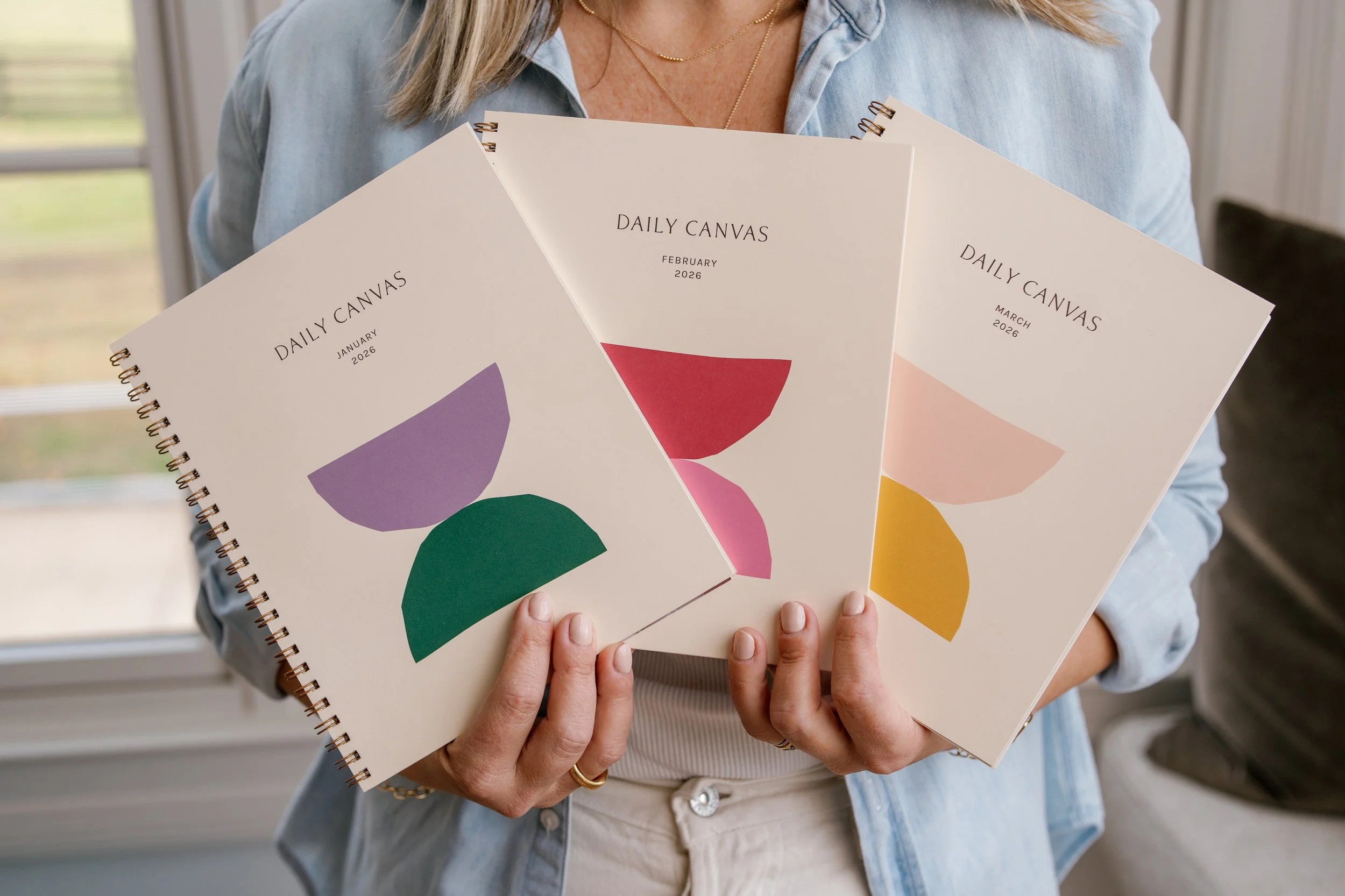 Woman holding a set of Daily Canvas spiral bound monthly planners, fanned out to showcase minimalist covers with soft neutral backgrounds and colorful geometric half circle designs.