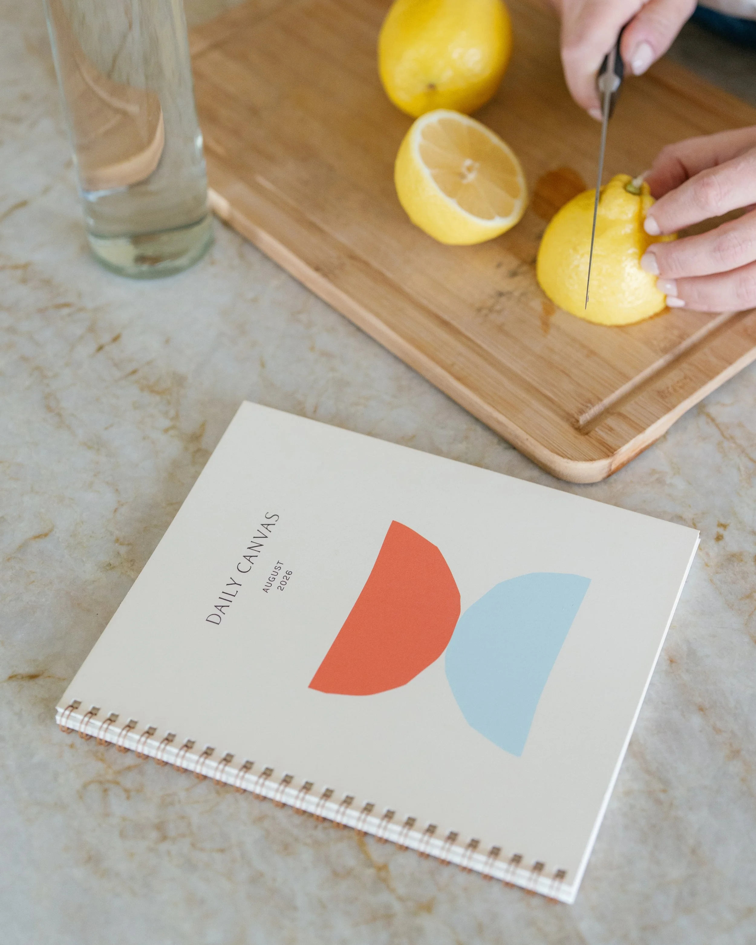 Daily Canvas spiral planner with a coral and blue geometric cover resting on a marble kitchen counter next to fresh lemons.