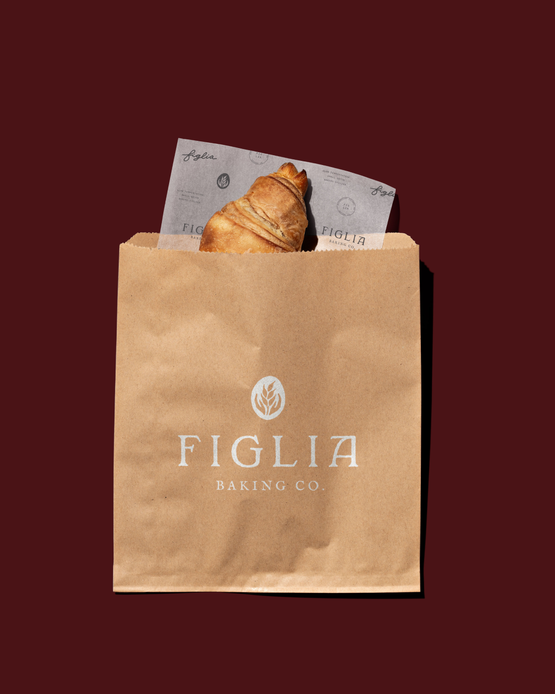 A brown paper bag printed with the Figlia Baking Co. logo sits against a dark maroon background. A croissant peeks out from the top, wrapped in branded paper patterned with the Figlia logo and circular wheat emblems.
