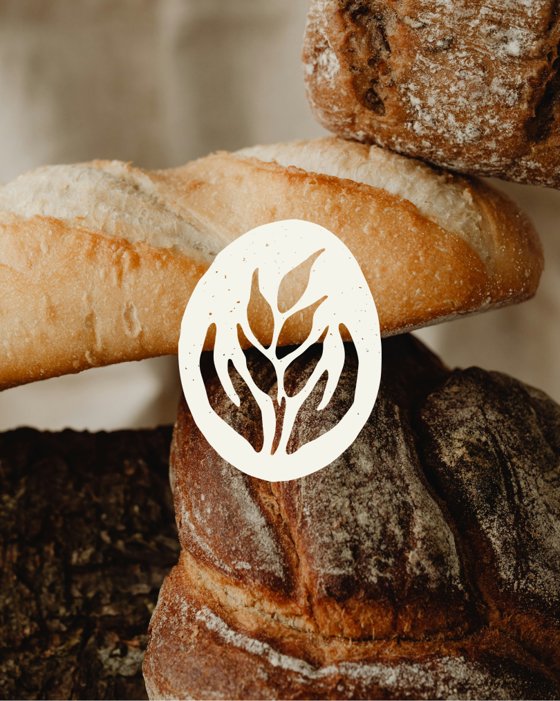 A stack of rustic baguettes and round loaves dusted with flour. A white wheat emblem enclosed in an oval is centered over the bread.