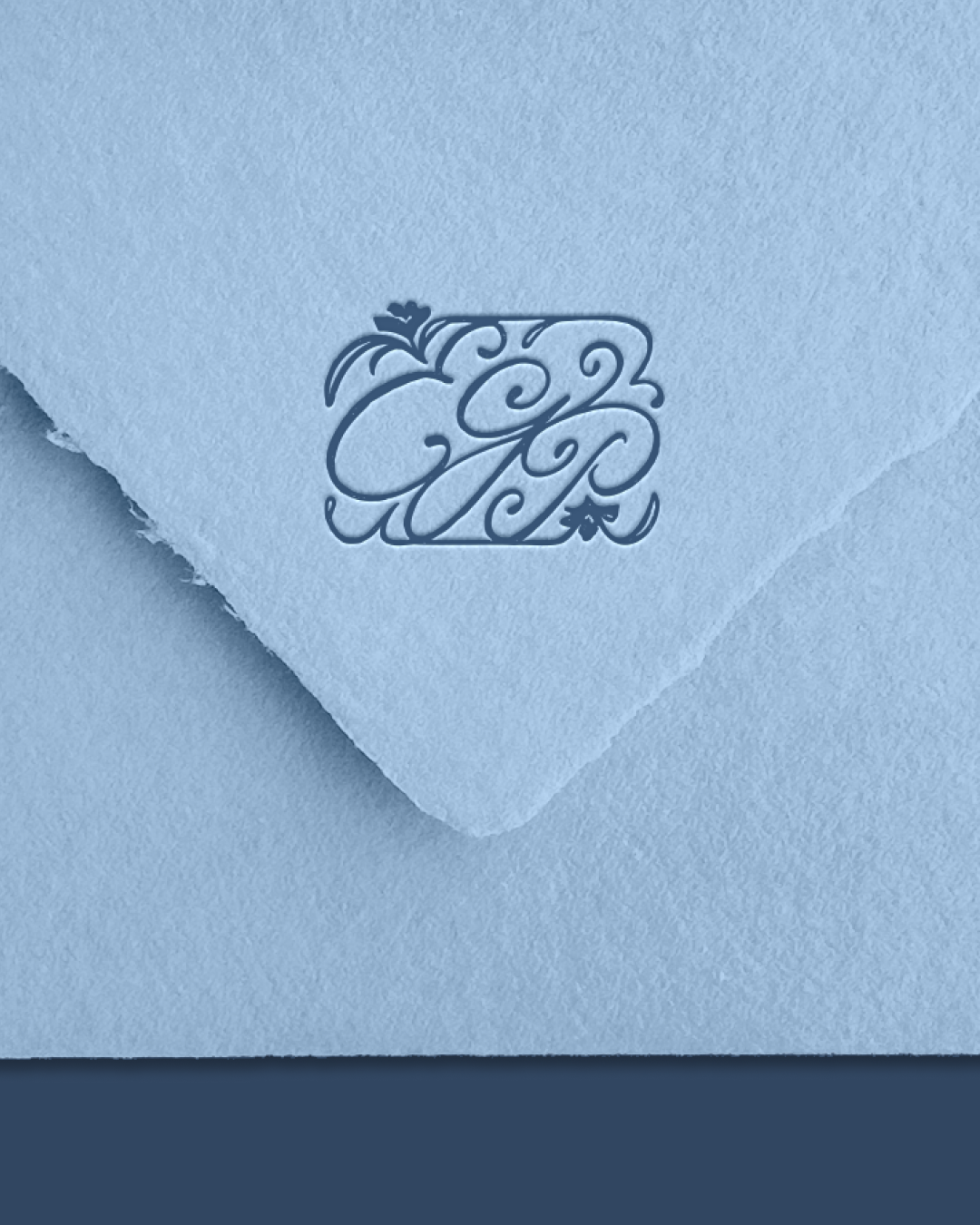 A close-up of a textured blue envelope with its flap partially open, showcasing an embossed decorative monogram made of ornate swirls and flourishes.